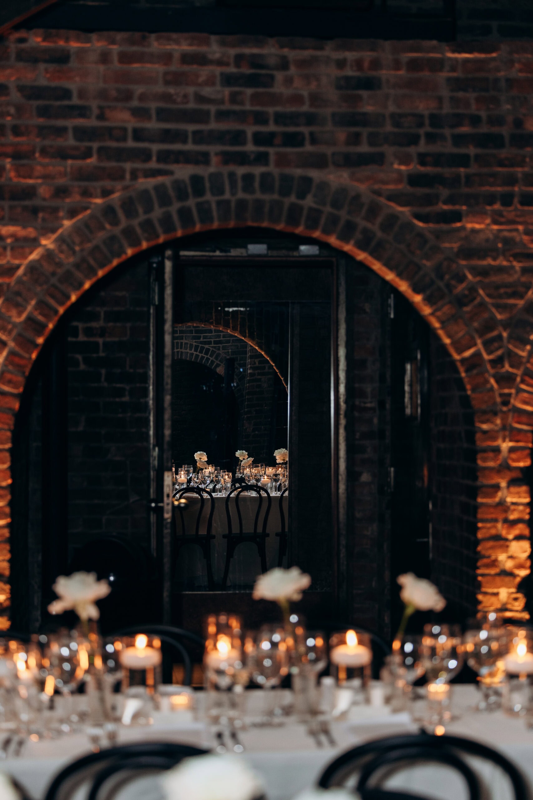 Candlelit wedding reception table seen through brick arches at The Foundry, with bentwood chairs, white florals, and glassware in a dimly lit interior.