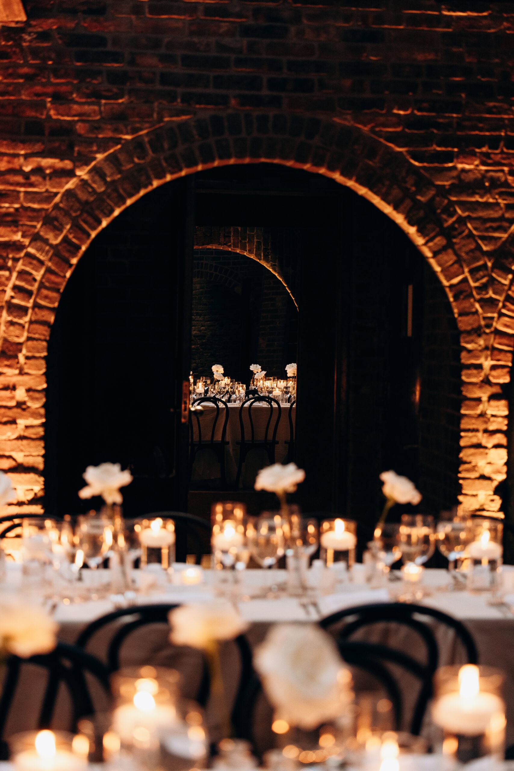 Candlelit wedding reception at The Foundry framed by brick archways, with white floral centerpieces, bentwood chairs, and layered tables glowing in low light.