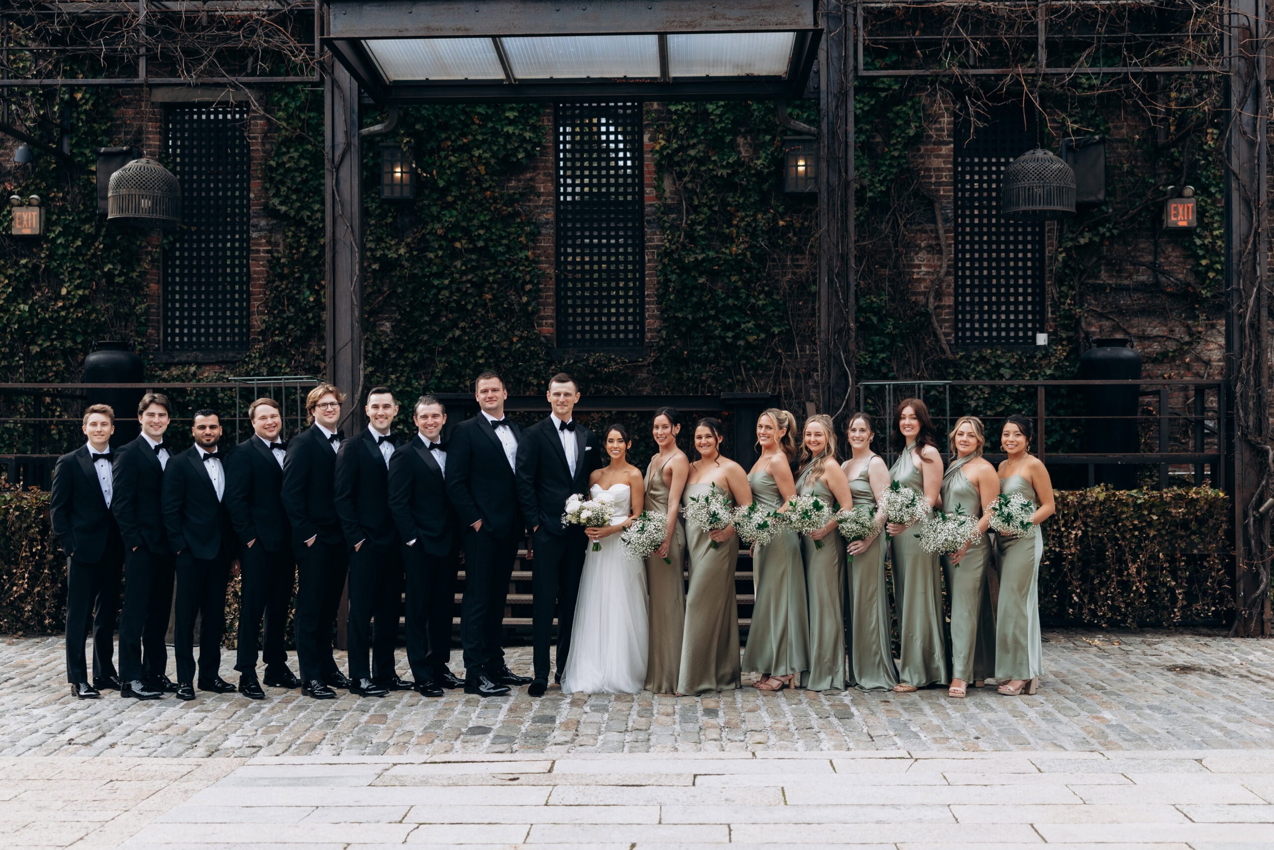 Wedding party portrait at The Foundry with the couple centered between groomsmen in black tuxedos and bridesmaids in sage satin dresses holding white bouquets.