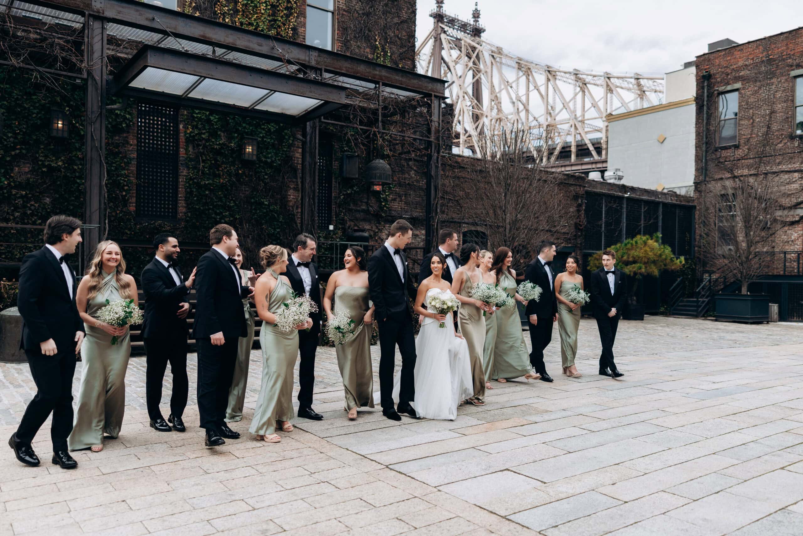Wedding party portrait in the courtyard at The Foundry with the bride in white, attendants in sage satin and black tuxedos, and the Queensboro Bridge behind.