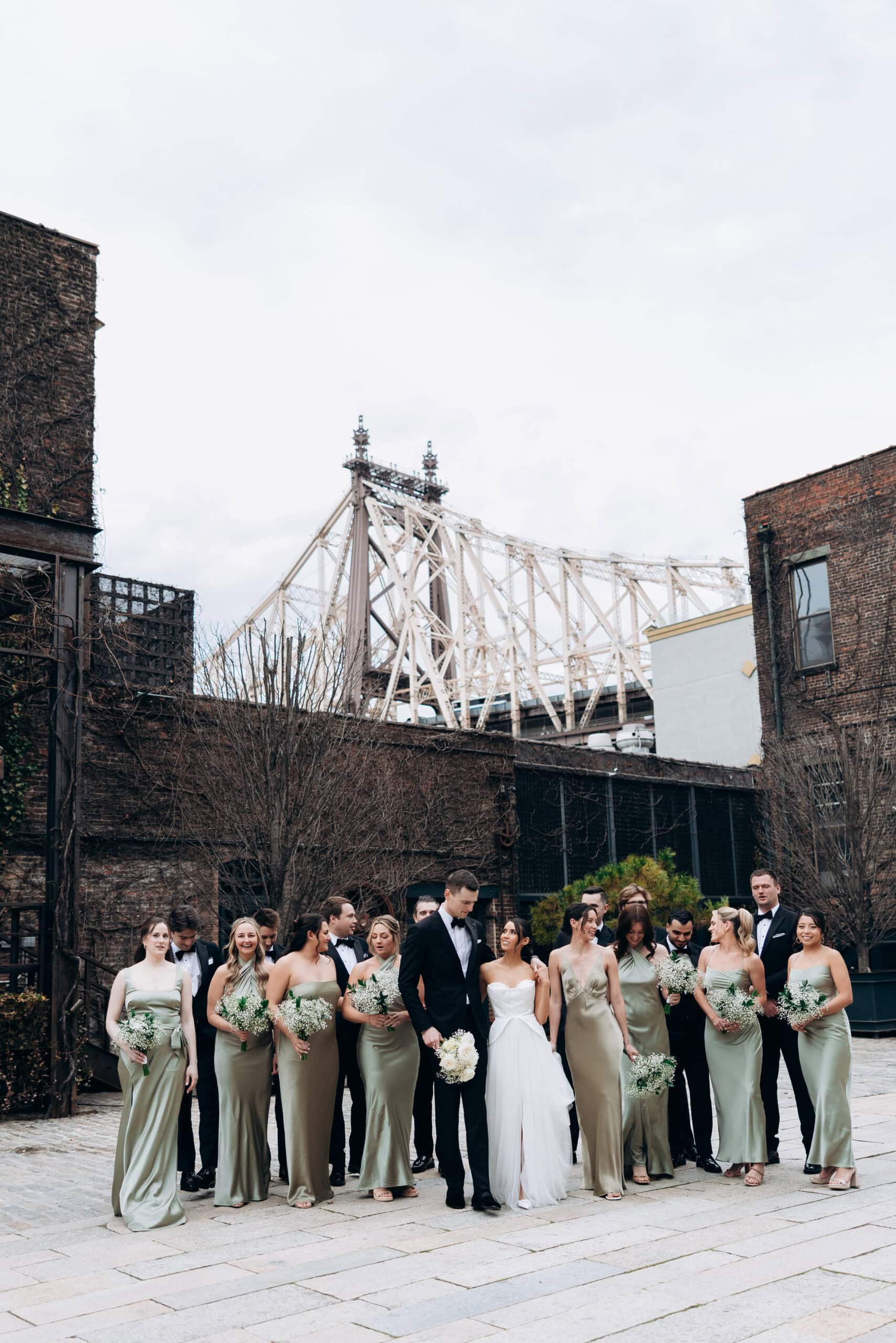 Wedding party in sage gowns and black tuxedos in an industrial courtyard with the Queensboro Bridge behind them in New York City, photographed March 27, 2026.
