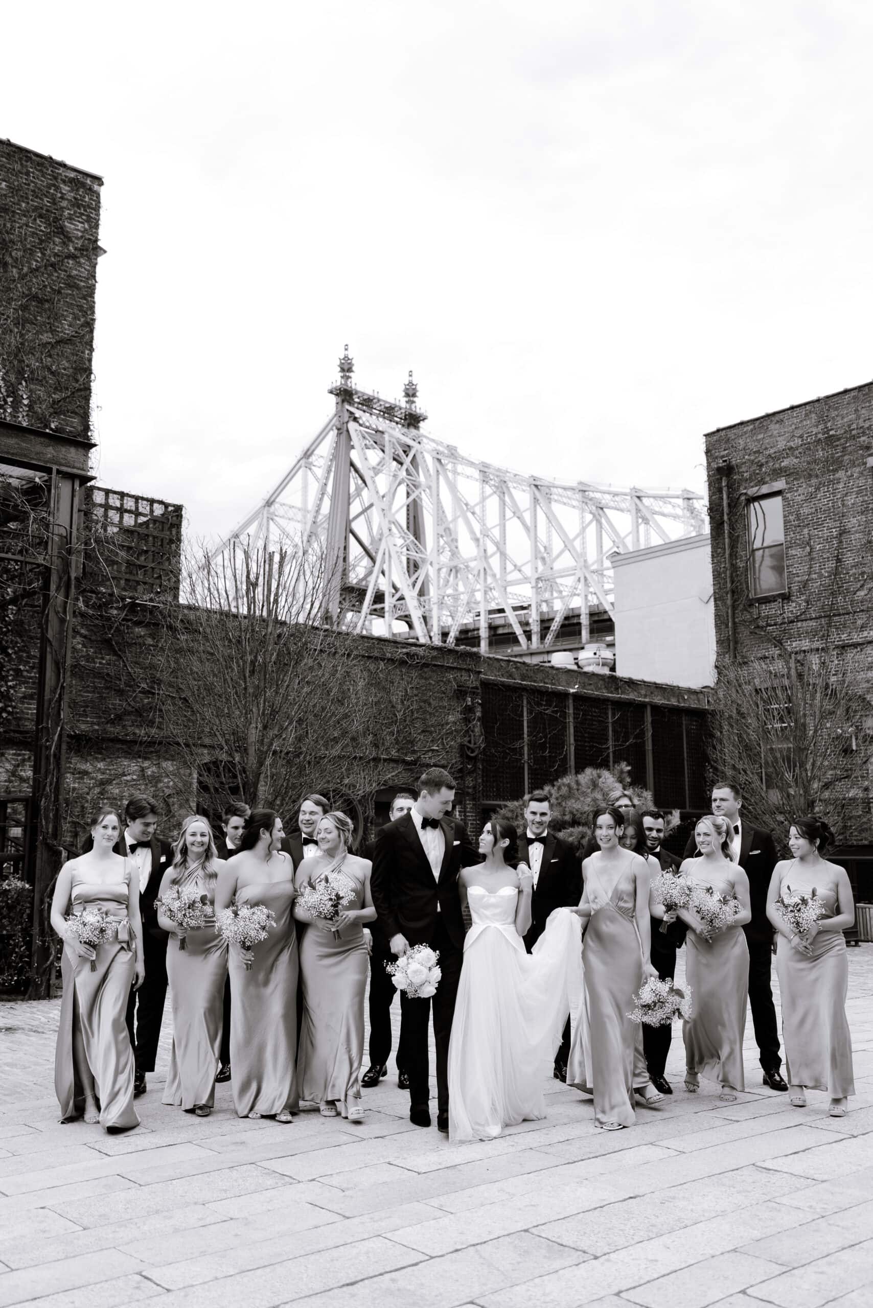 Black-and-white wedding party portrait at The Foundry courtyard, with the couple and attendants walking beneath the Queensboro Bridge framed by brick buildings.
