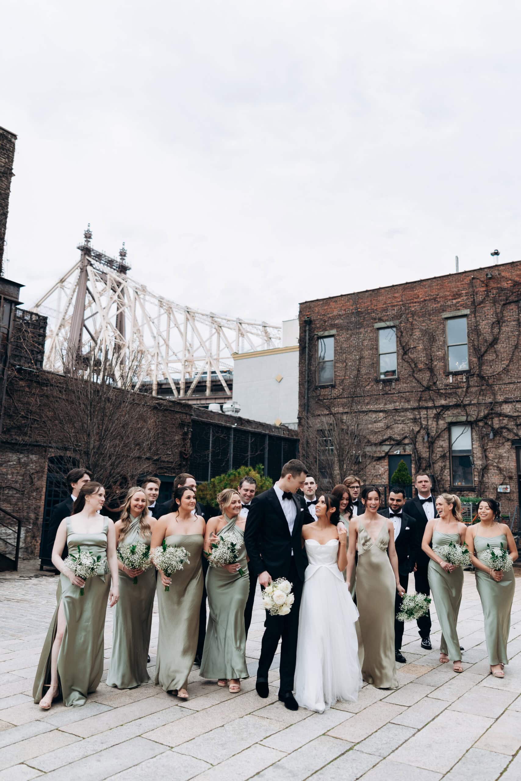 Wedding party walking in The Foundry courtyard beneath the Queensboro Bridge, with sage satin dresses, black tuxedos, brick facades, and an overcast sky.