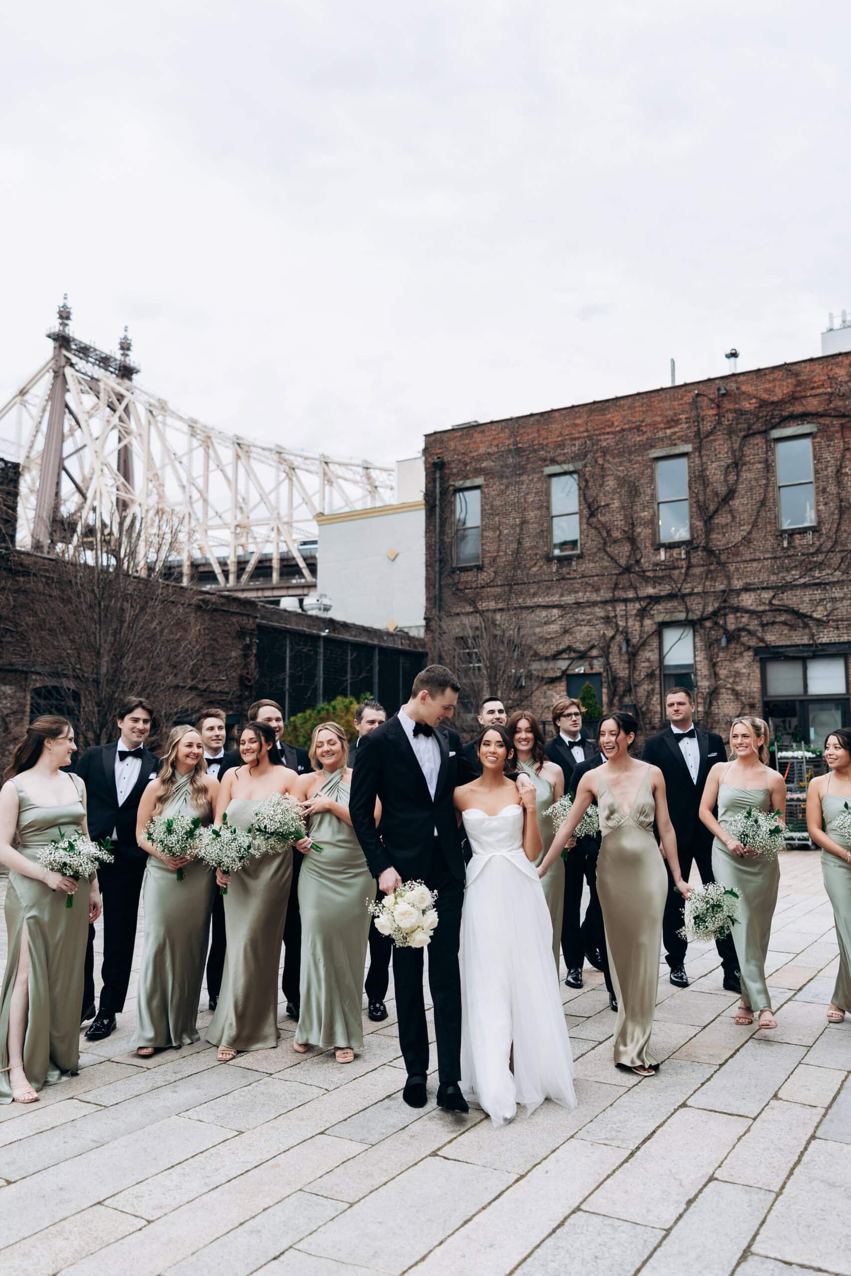 Bride and groom walk with their wedding party in the courtyard at The Foundry, with brick buildings and a large steel bridge in the background.