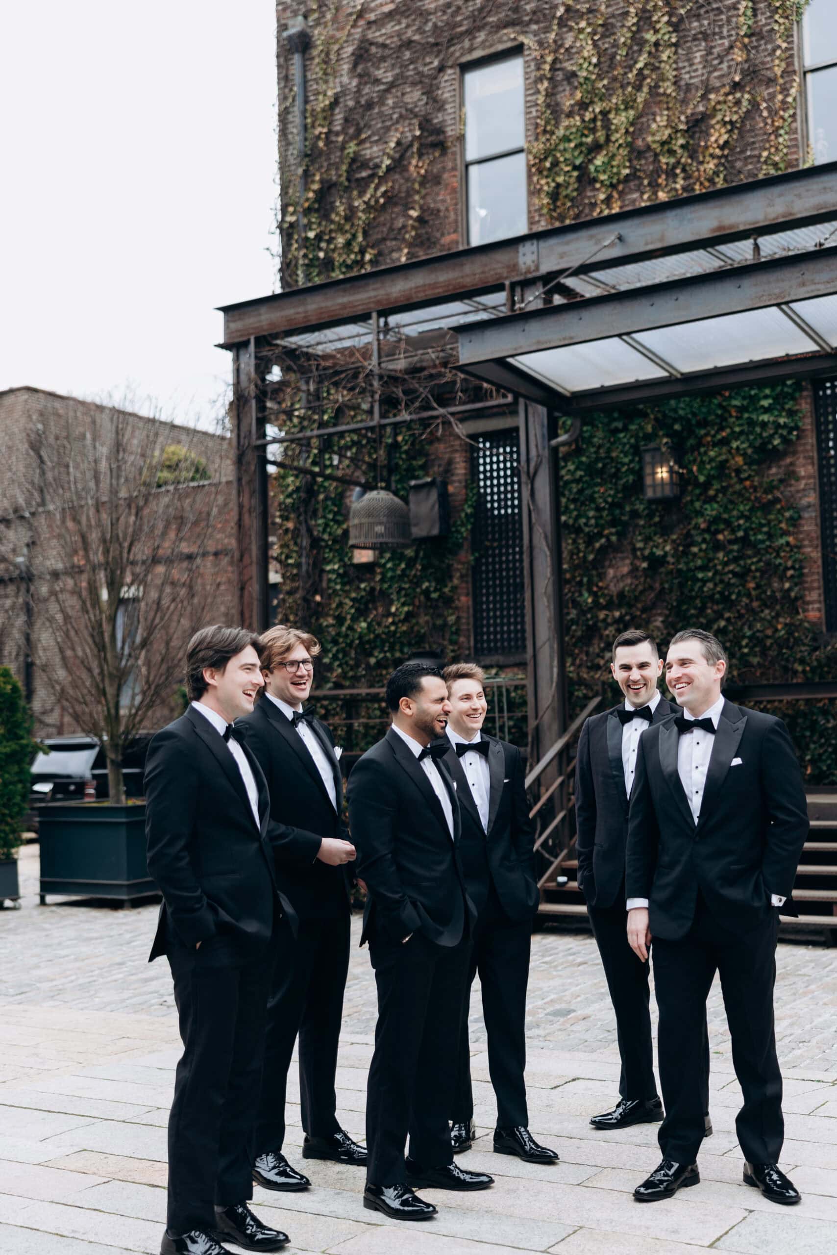Groomsmen in black tuxedos laughing together in the ivy-covered courtyard at The Foundry wedding venue in New York City.