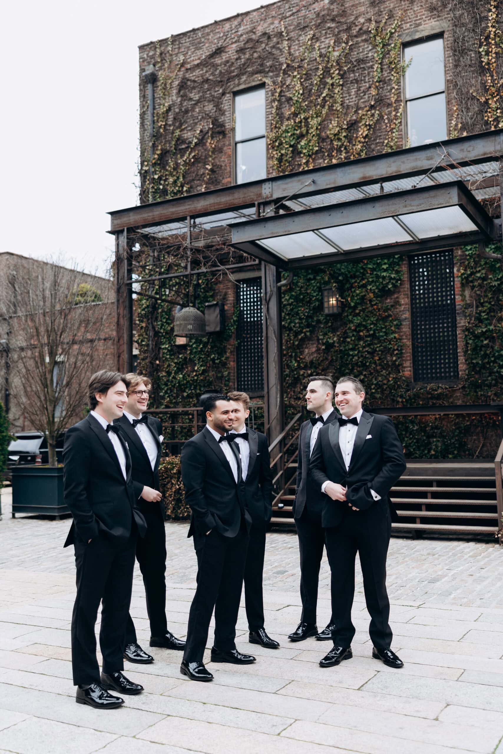 Five men in black tuxedos stand together in a stone courtyard outside The Foundry, with ivy-covered brick walls and an industrial canopy behind them.