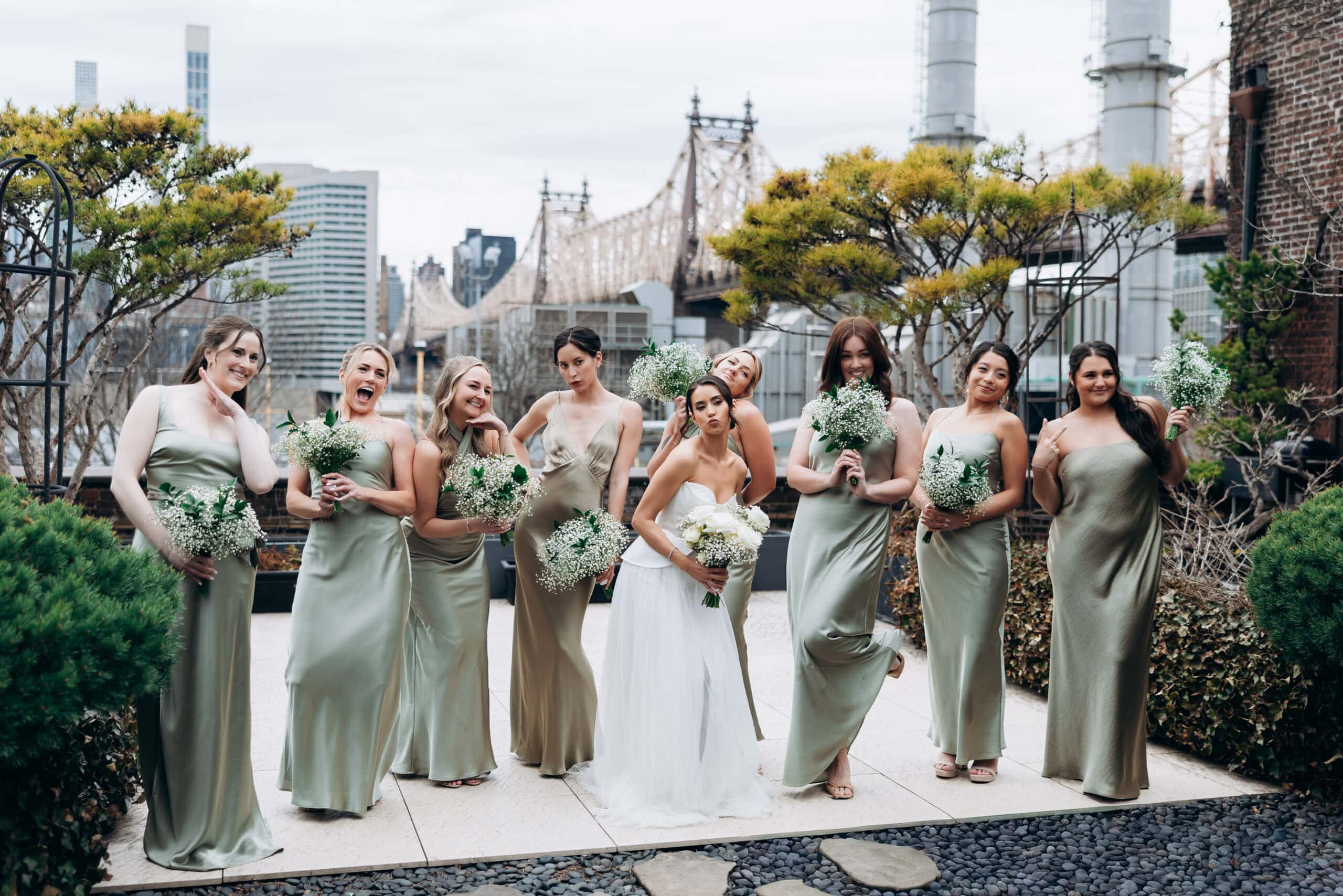 Bride in a white gown posing with eight bridesmaids in sage satin dresses holding white bouquets on the terrace at The Foundry, with the Queensboro Bridge behind them.