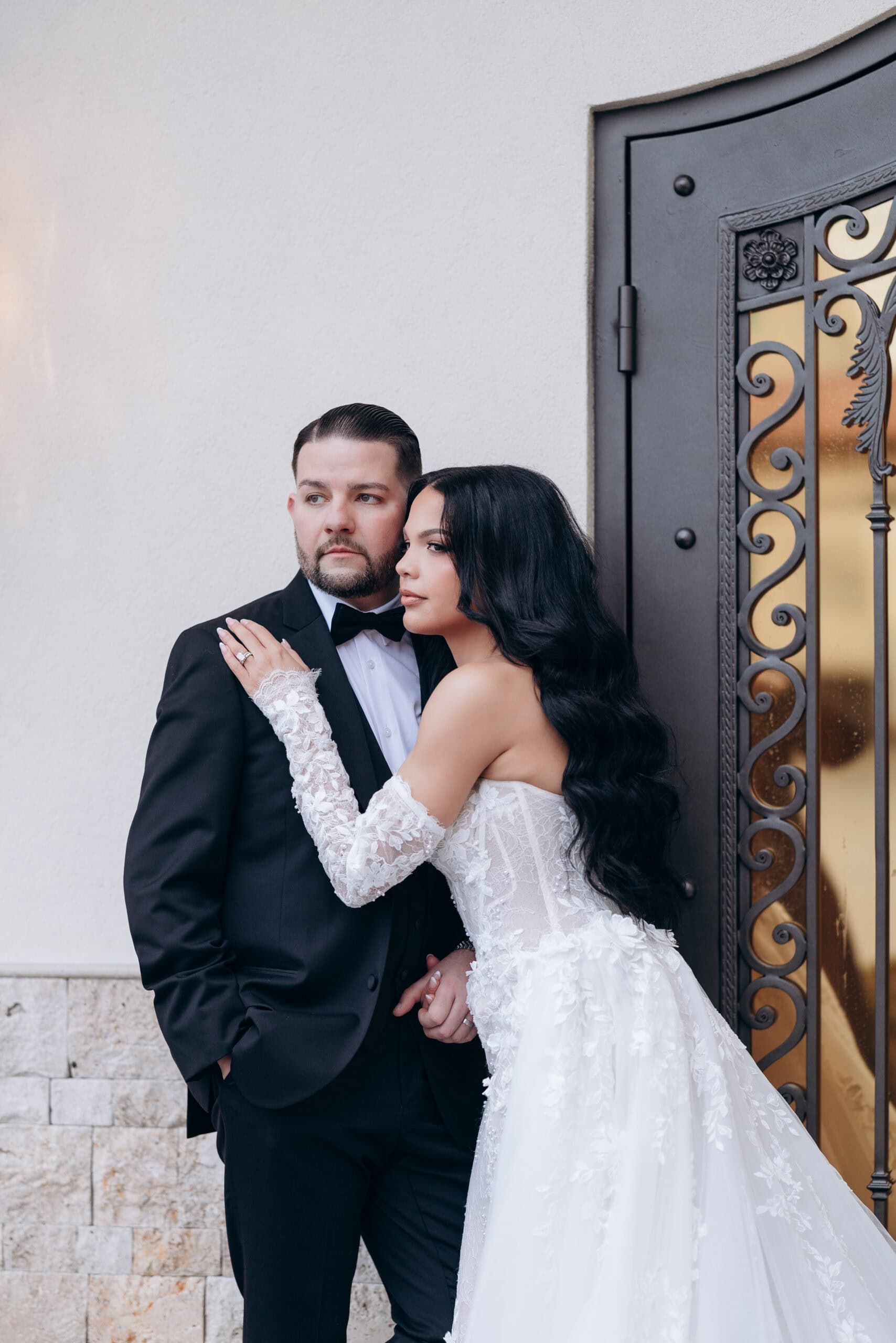 Bride in lace sleeve wedding gown embraces groom in black tuxedo near ornate iron door with golden glass, against textured stone and plaster wall at The Grand.
