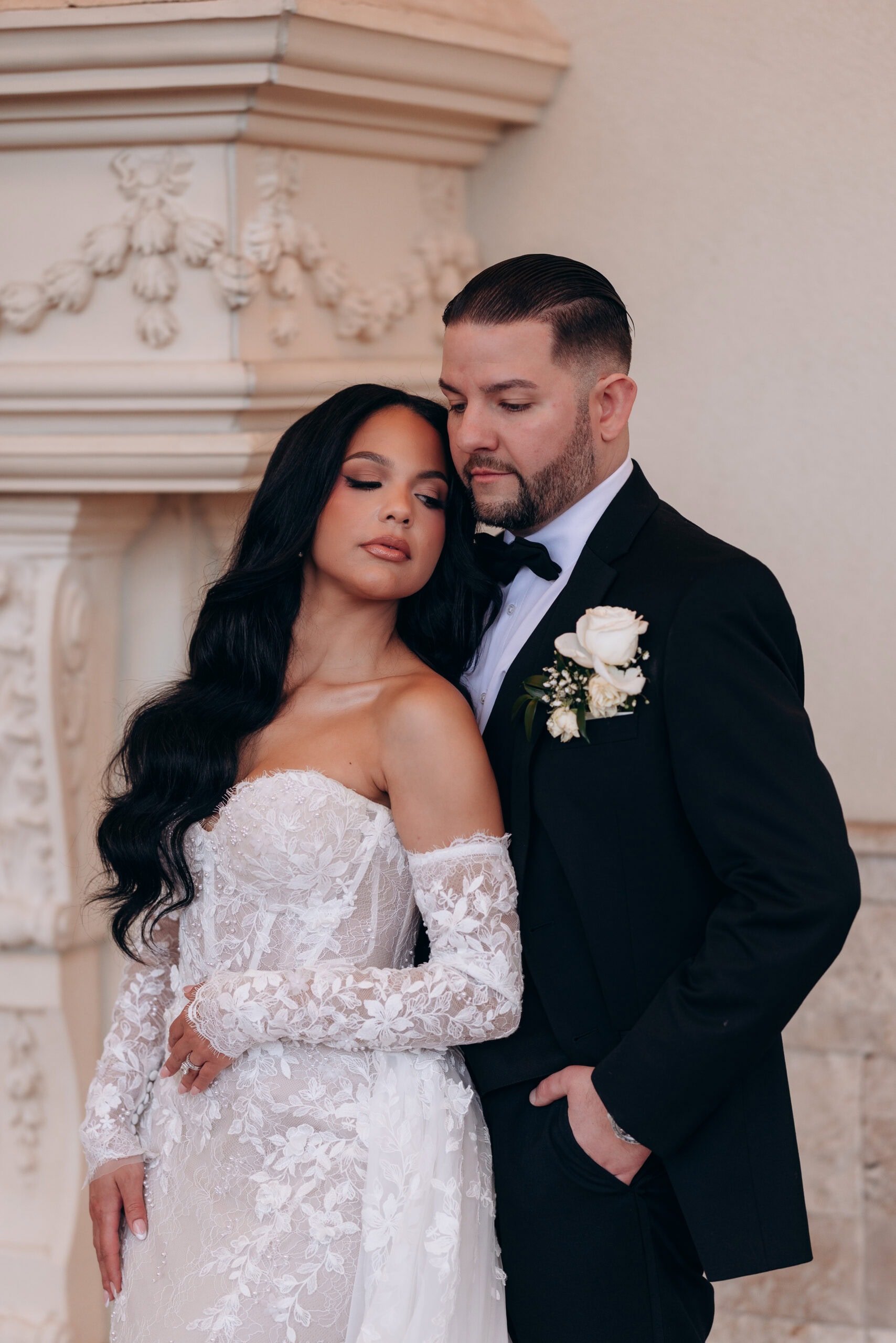 Bride in lace off-shoulder wedding dress resting head on groom's shoulder, groom in black tuxedo with white rose boutonniere, standing near ornate molding at The Grand.