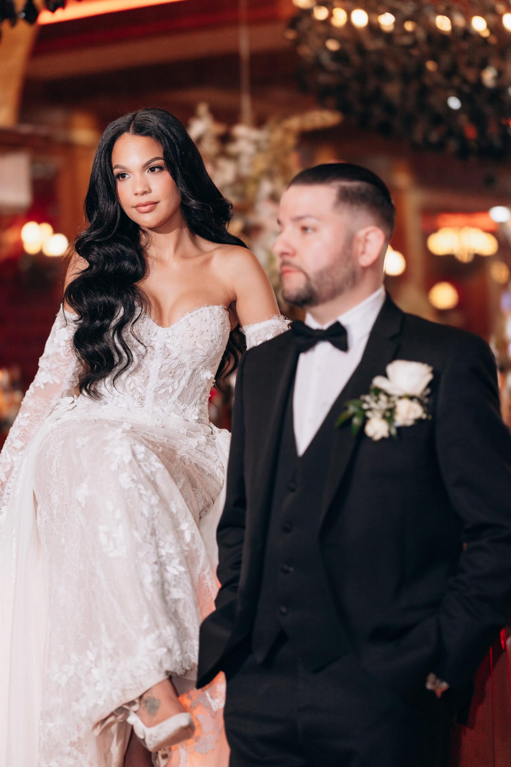 Bride in intricate lace wedding dress with long dark hair, seated and looking away; groom in black tuxedo with white rose boutonniere stands in front, softly blurred, warm indoor lighting.