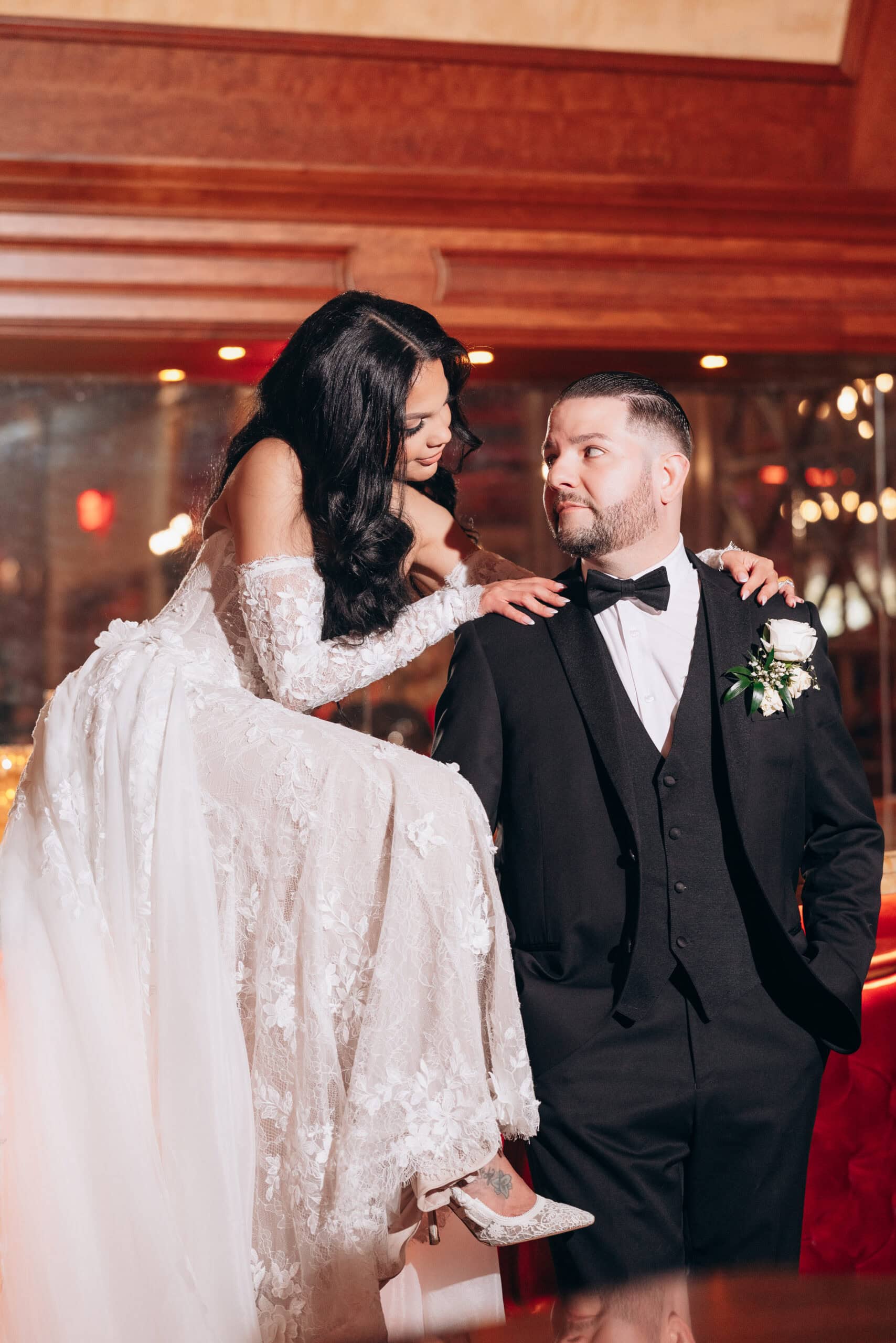 Bride in lace wedding gown and embroidered heels looks lovingly at groom in black tuxedo with white rose boutonniere inside a venue with wood paneling.