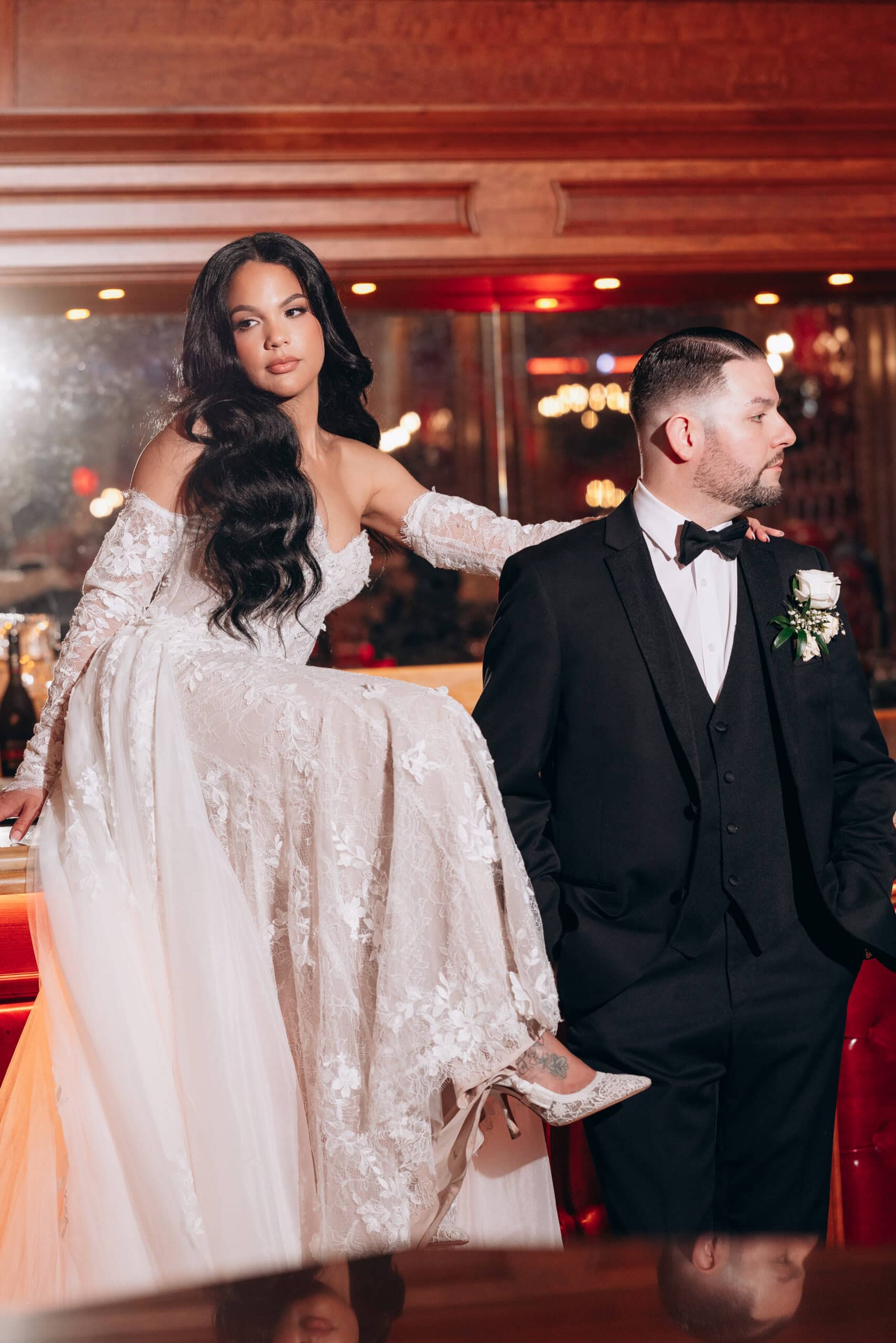Bride in detailed lace wedding dress with long dark hair seated beside groom in black tuxedo with white rose boutonniere, indoor setting with warm wood paneling and soft lighting.
