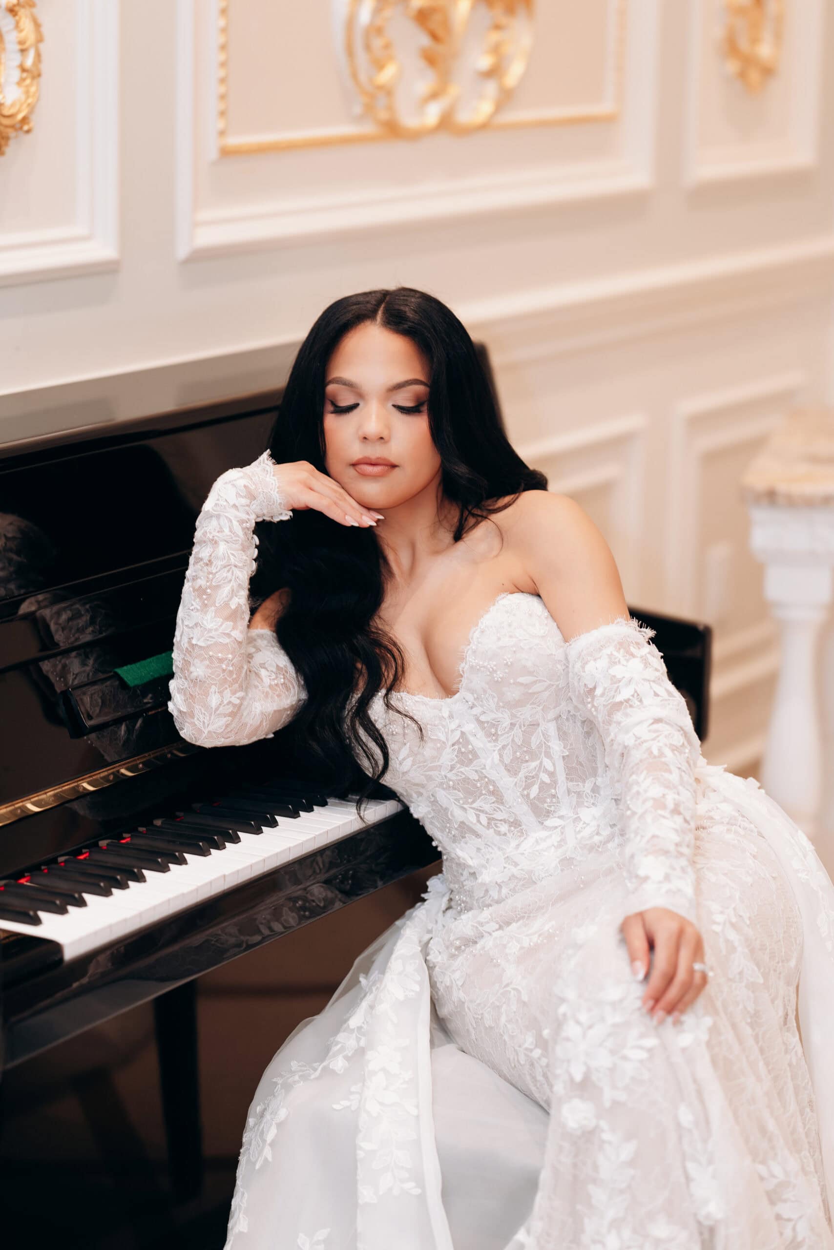 Bride in detailed lace wedding gown resting her head on her hand while seated at a black piano in an opulent room with gold-accented white walls.