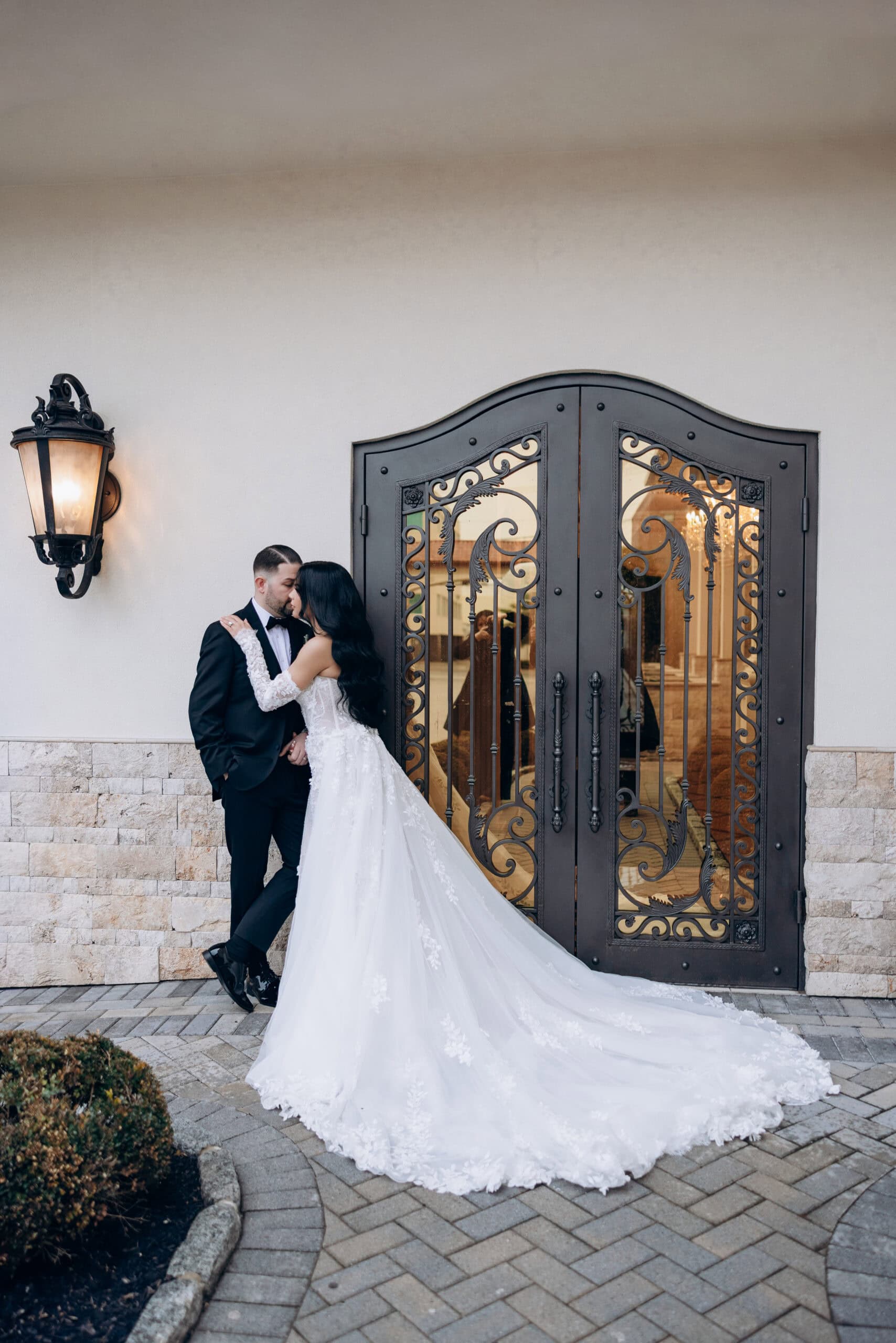 Bride in lace wedding gown embraces groom in black tuxedo by ornate wrought iron doors and stone wall at The Grand venue.