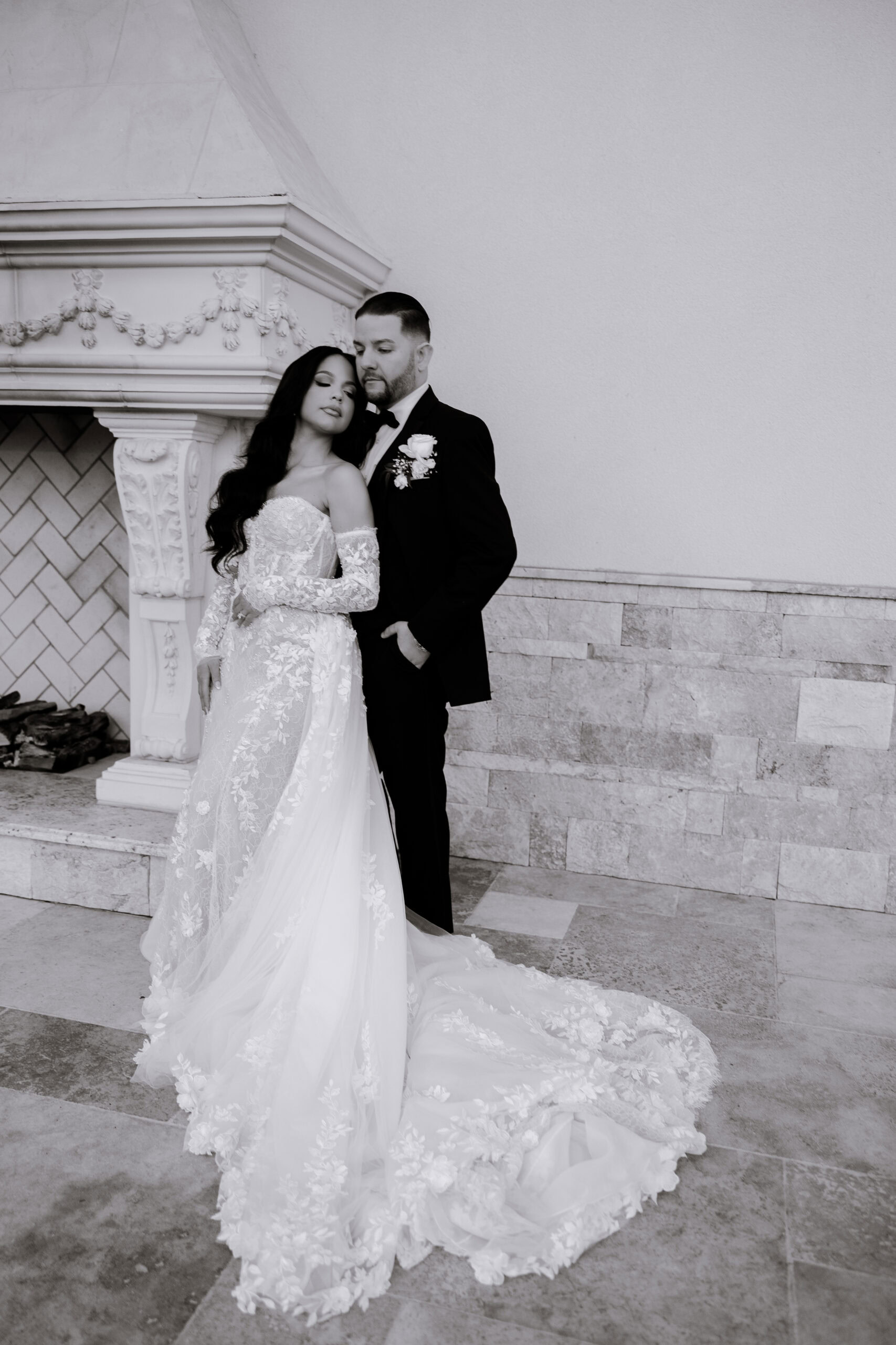 Bride in detailed lace wedding gown and groom in black tuxedo stand close by an ornate stone fireplace, showcasing elegant textures and intimate pose.