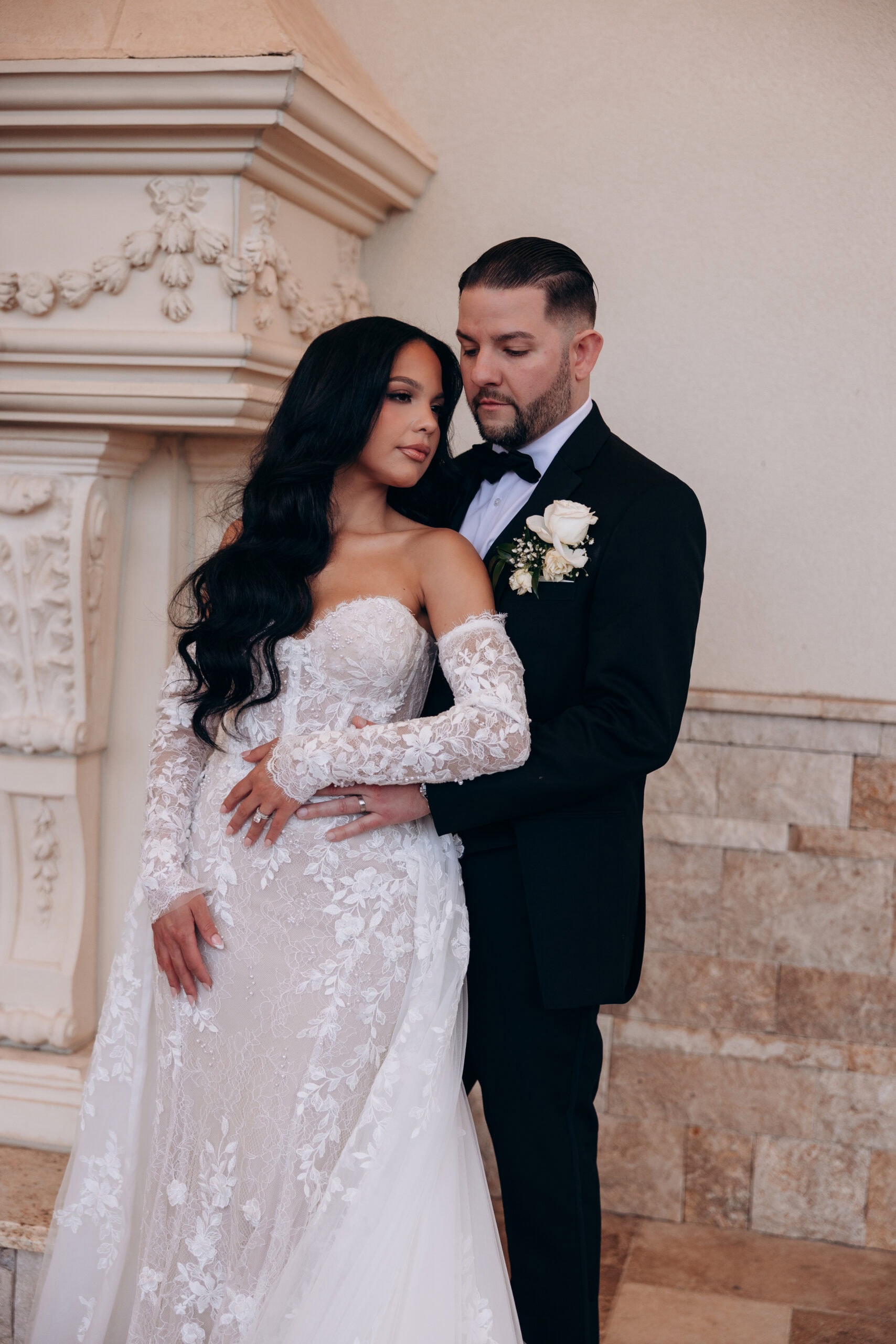 Bride in intricate lace wedding gown with long sleeves embraced by groom in black tuxedo with white rose boutonniere, standing near ornate stone wall at The Grand.