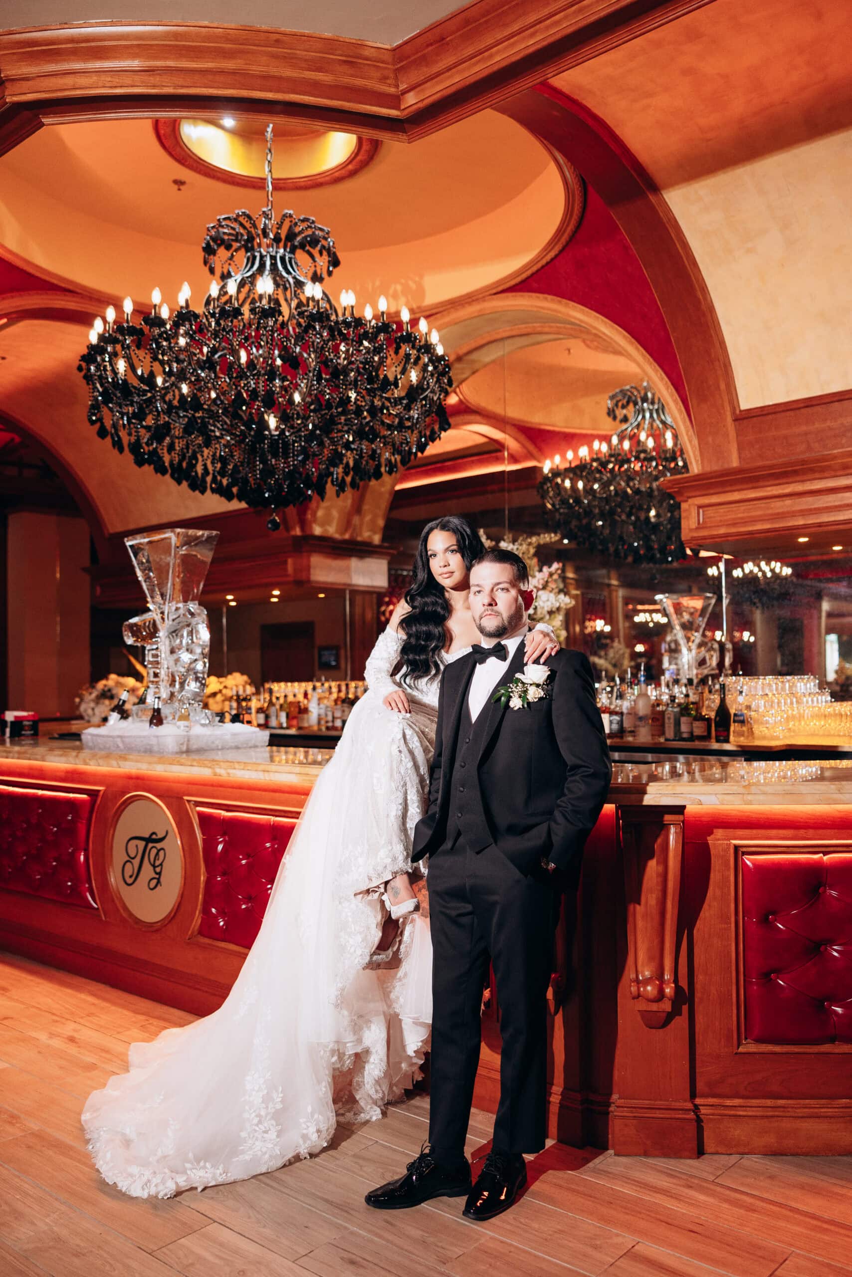 Bride in lace wedding gown and groom in black tuxedo standing in a luxurious bar with red leather seating and large black crystal chandelier overhead at The Grand.