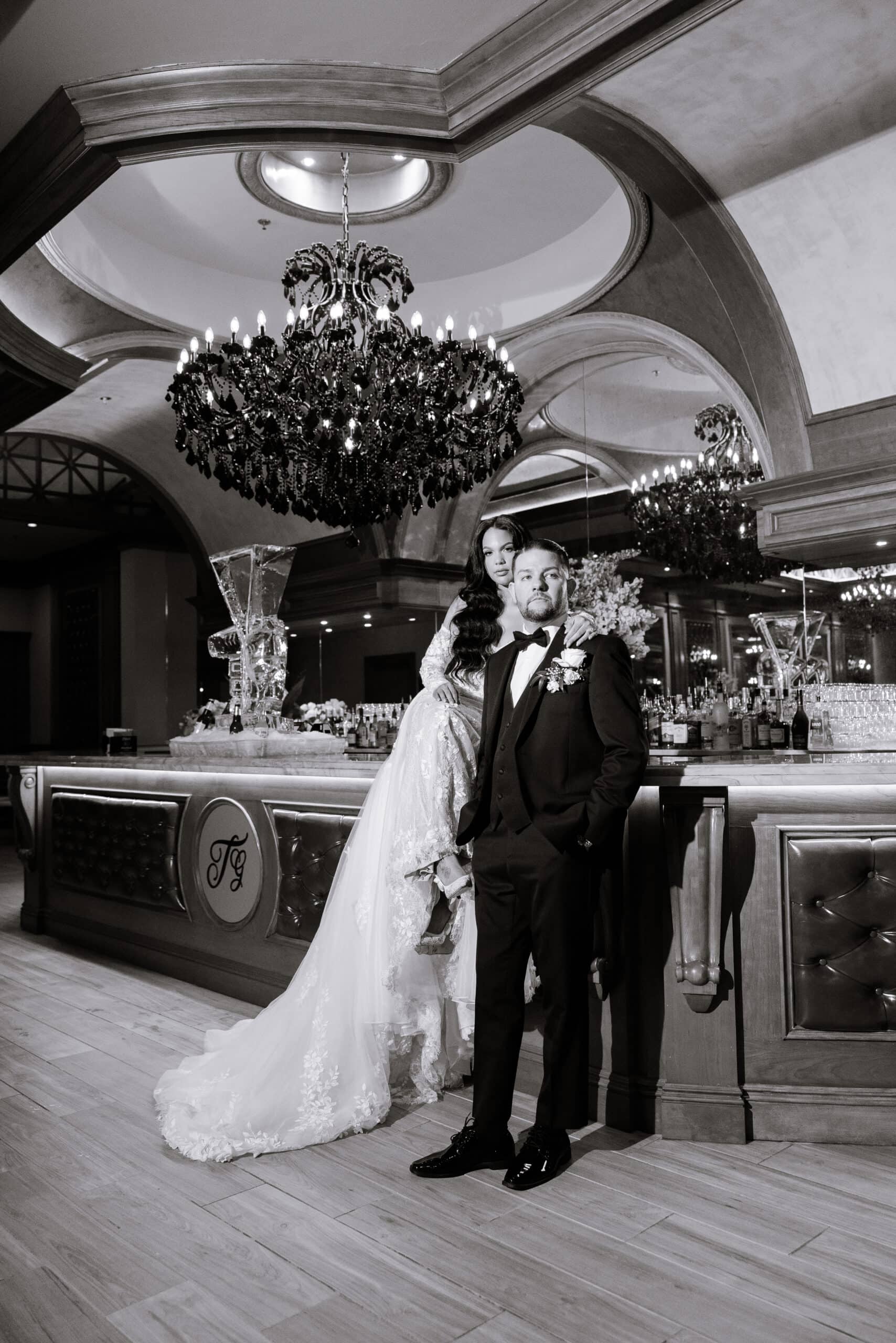 Black and white photo of a bride in lace gown and groom in tuxedo standing in an elegant bar with large chandelier and mirrored arches at The Grand.