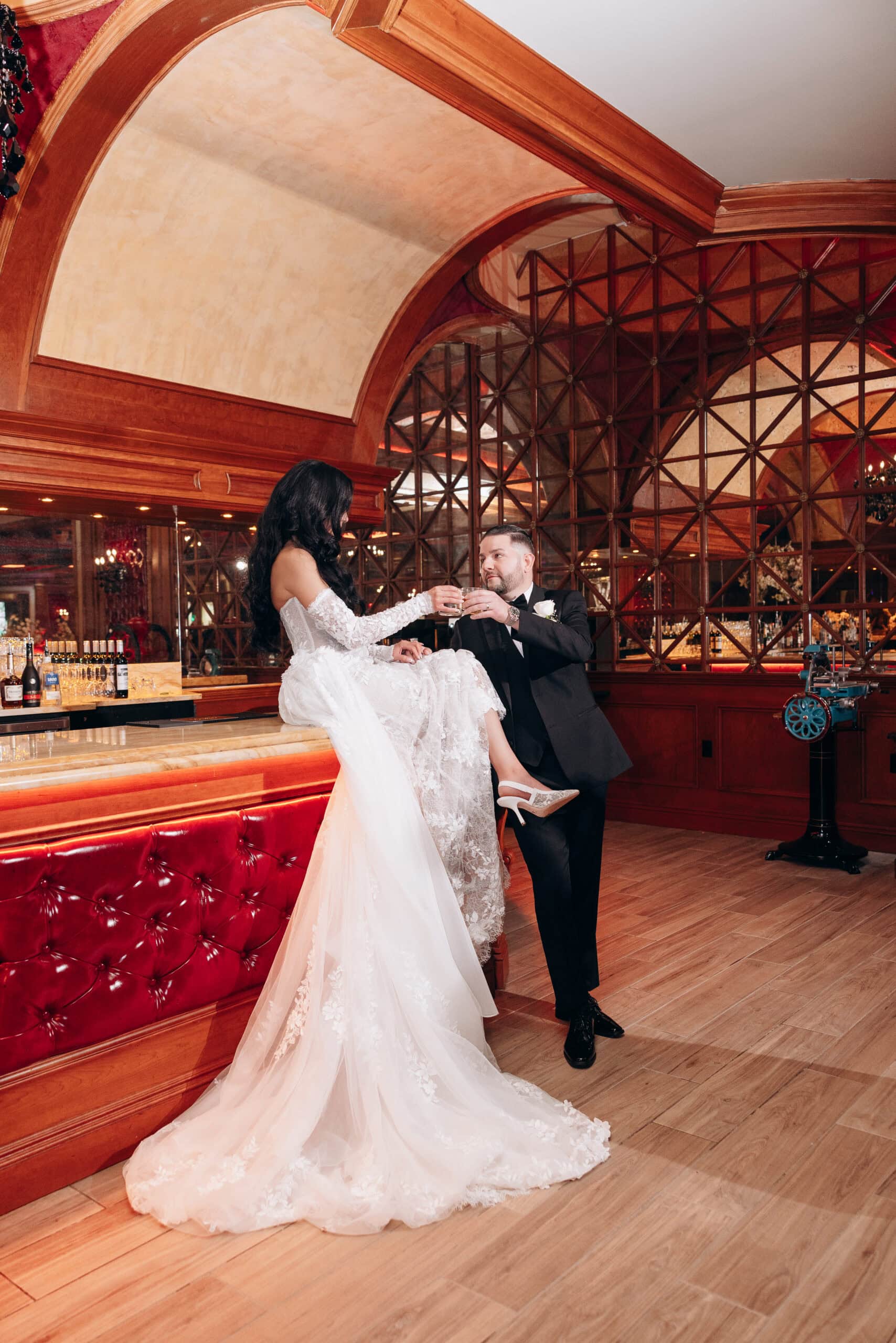 Bride in lace wedding dress sitting on marble bar counter toasting groom in black tuxedo inside a luxurious bar with wood arches and mirrored wall panels