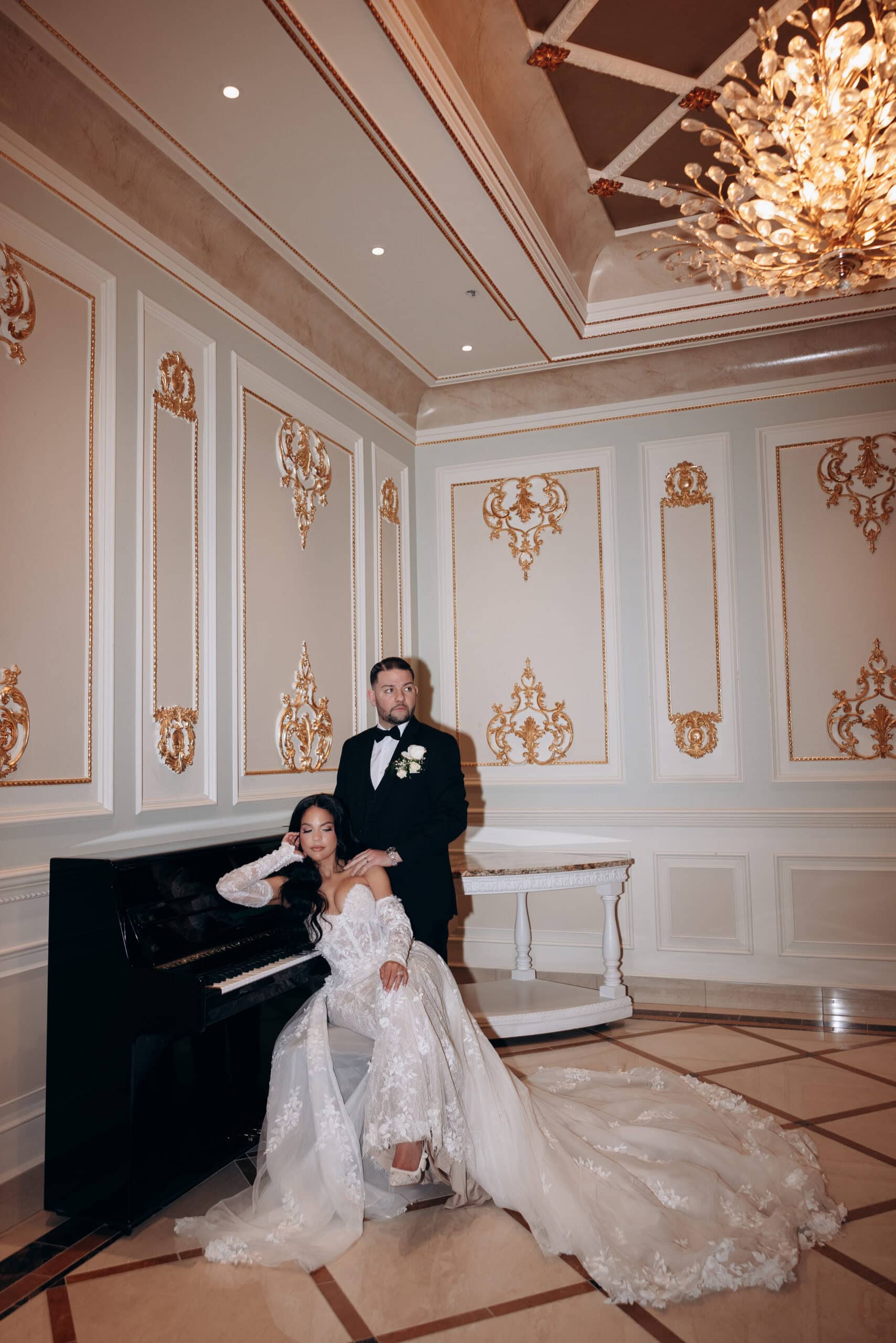Bride in lace wedding gown seated at black grand piano with groom in tuxedo standing behind her in an ornately decorated ballroom with gold accents and chandelier.