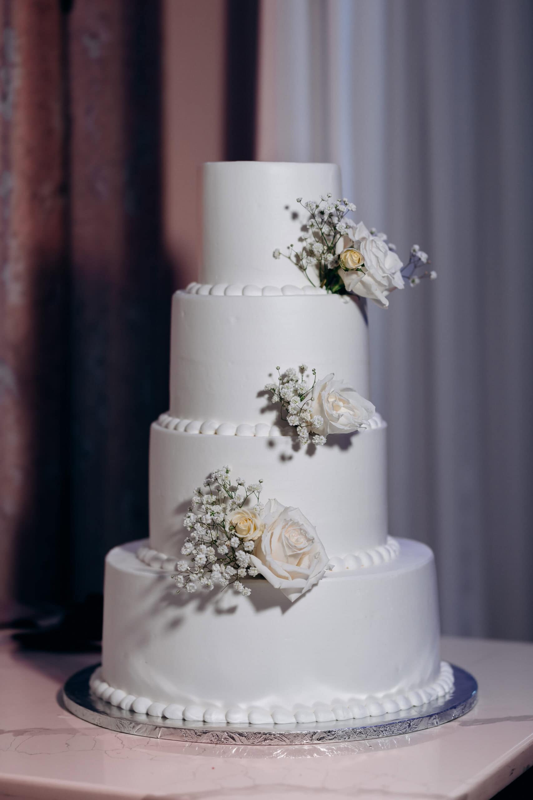 Four-tier white wedding cake decorated with white roses and baby's breath flowers, displayed on a marble table with a purple curtain background.