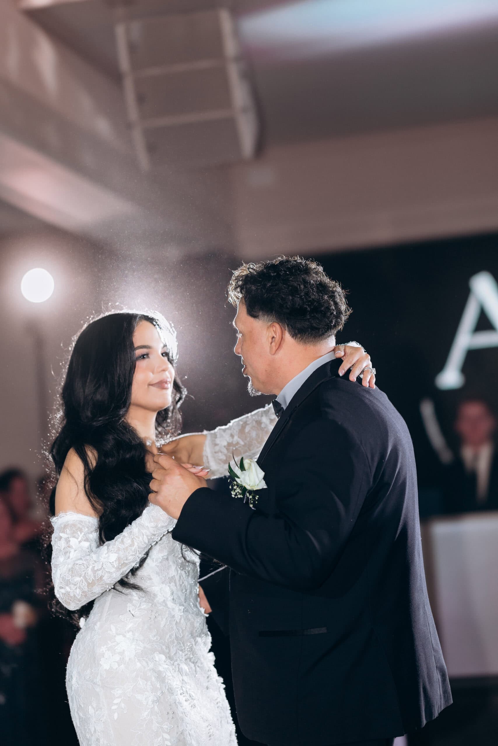 Bride in detailed lace wedding dress dancing with her father in a dark suit, softly backlit creating a halo effect around them.