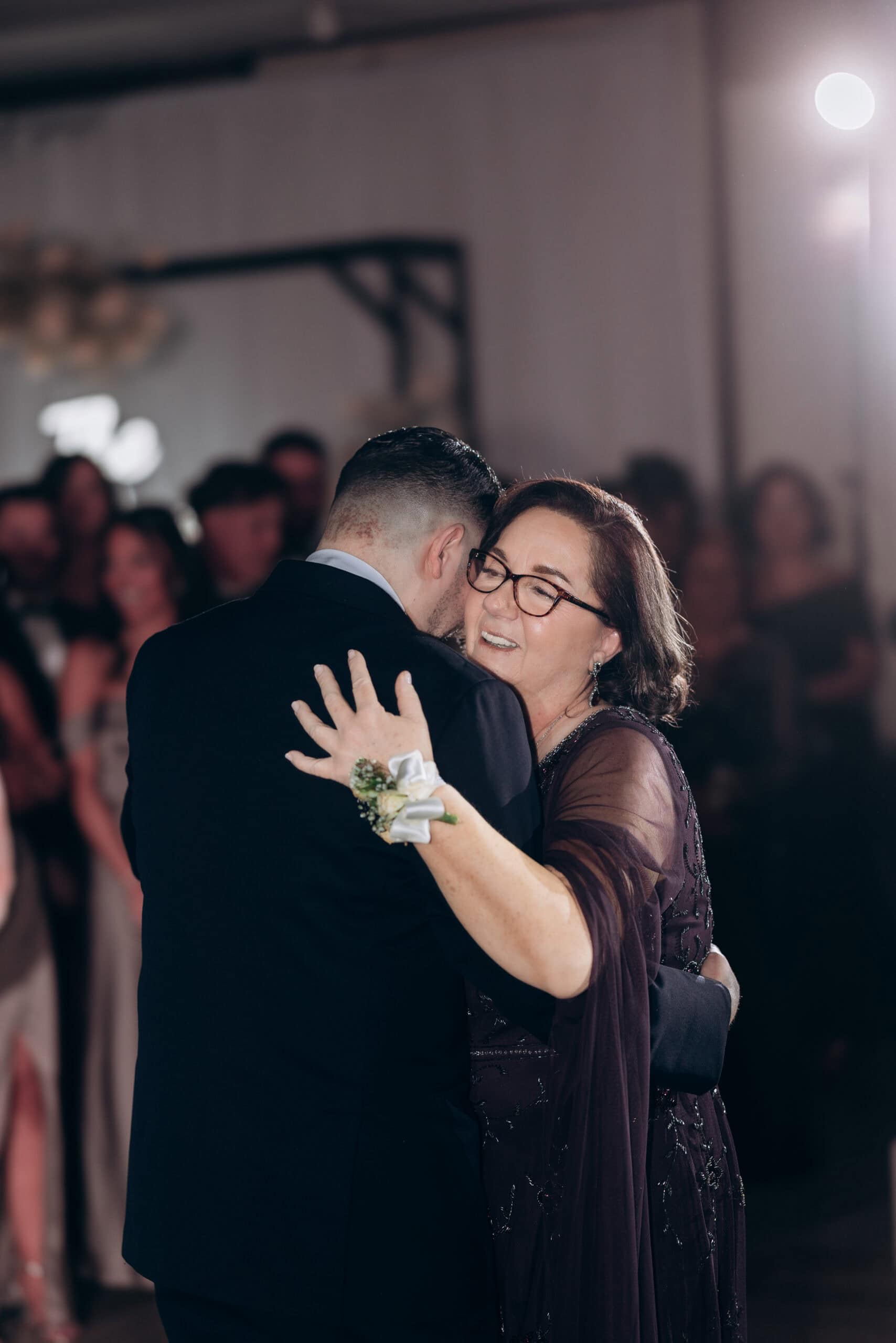 Groom in black tuxedo embraces his mother in a deep purple embroidered dress during a wedding dance, with blurred guests in the background.
