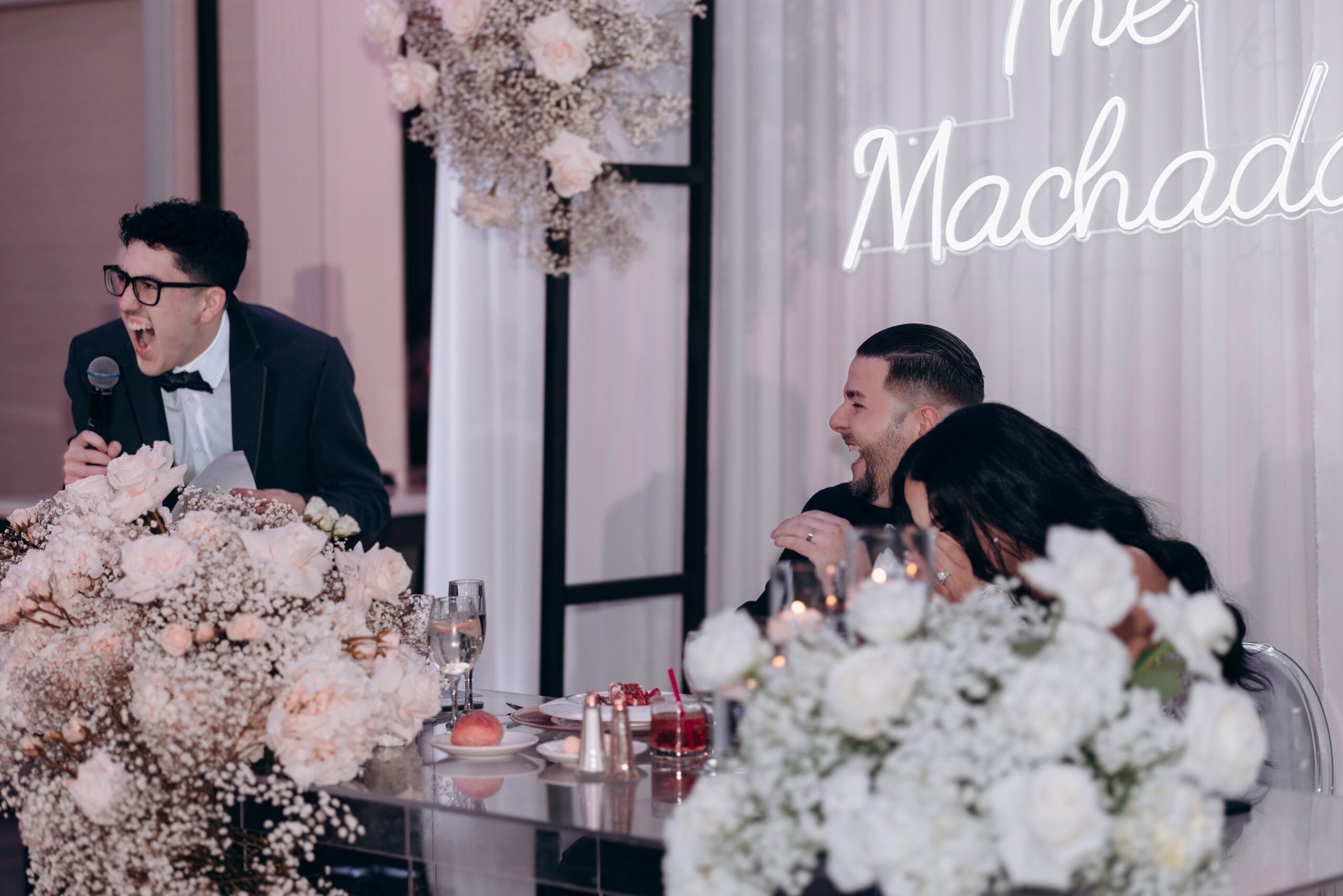 Wedding speech with a man in glasses laughing energetically holding a microphone, while the groom and bride laugh seated at a table adorned with white roses and baby's breath flowers.