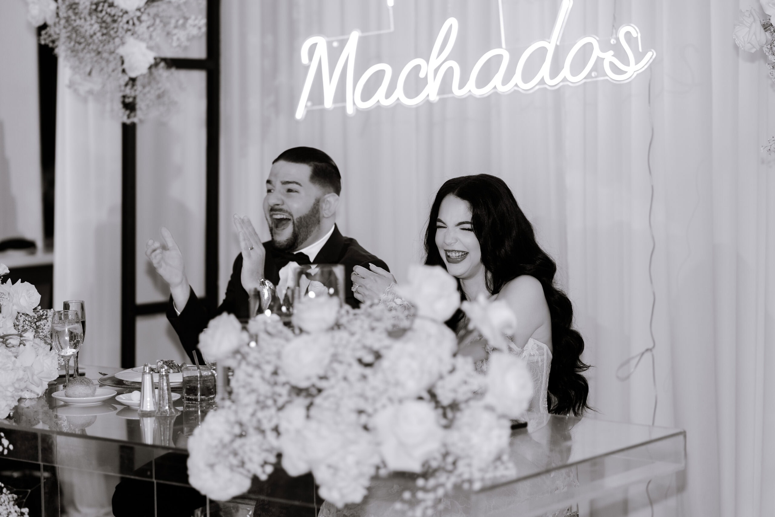 Bride and groom seated at a clear acrylic table adorned with white floral arrangements, laughing joyfully during their wedding reception under a glowing Machado's sign.