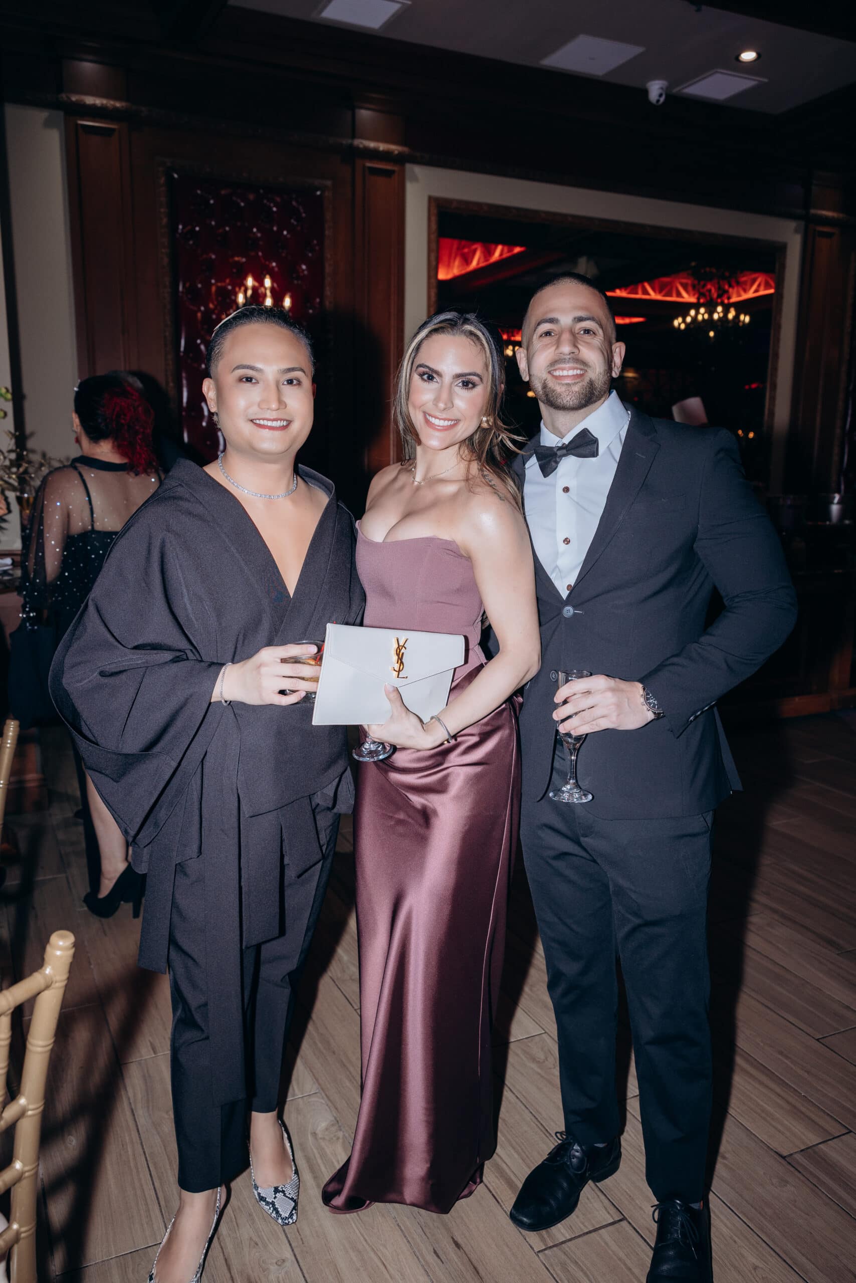 Three guests in formal eveningwear stand closely together indoors, holding drinks, with warm lighting and dark wood paneling in the background.