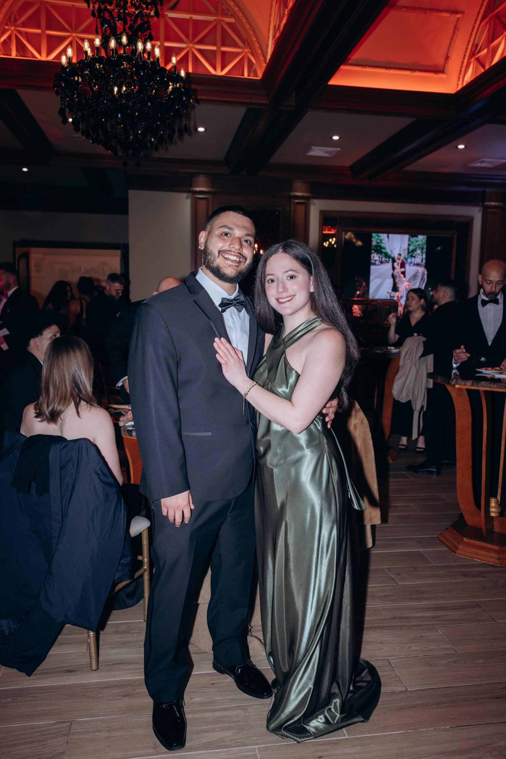 Man in black tuxedo and woman in olive satin gown smiling and embracing in an elegant ballroom with chandelier and wood accents, guests in background.