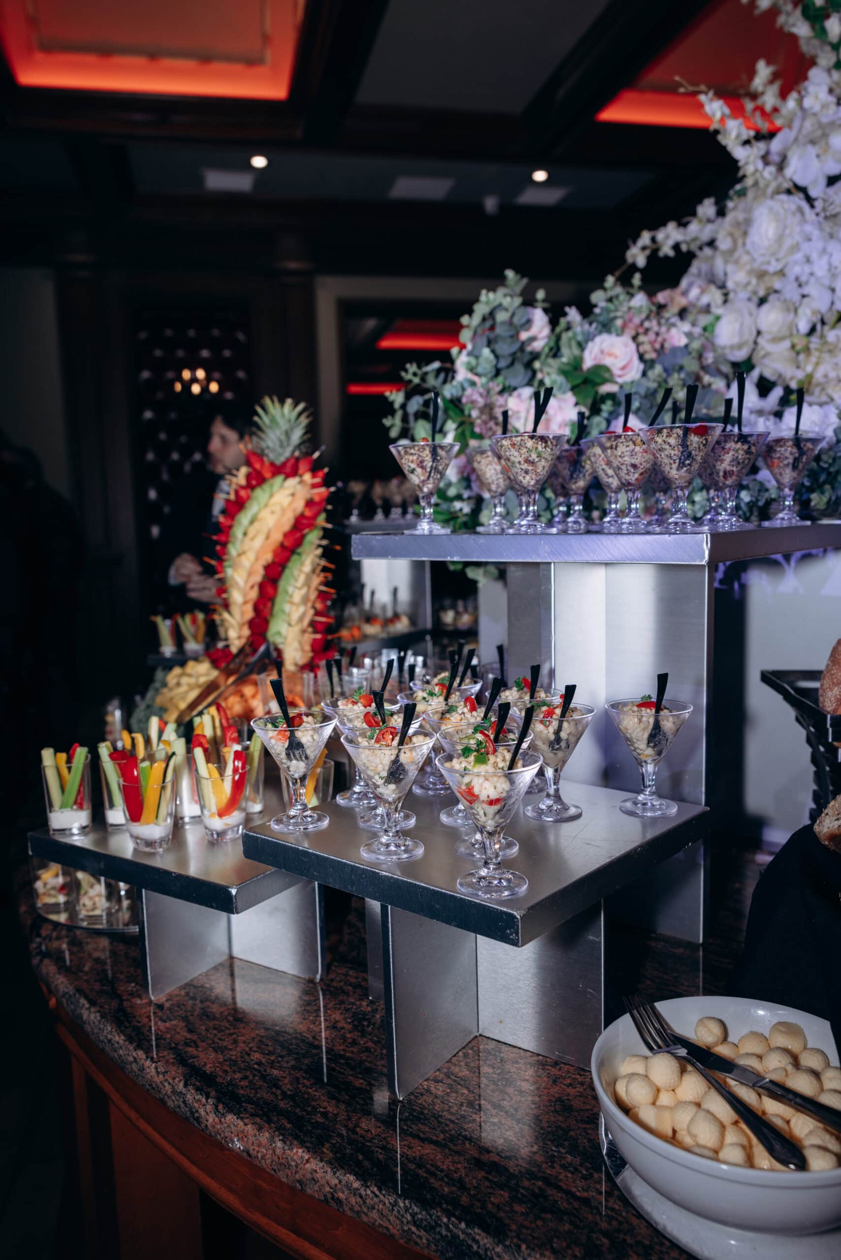Close-up of elegant hors d'oeuvres in martini glasses and vegetable sticks on a granite countertop with floral arrangements in a dimly lit luxury wedding venue.