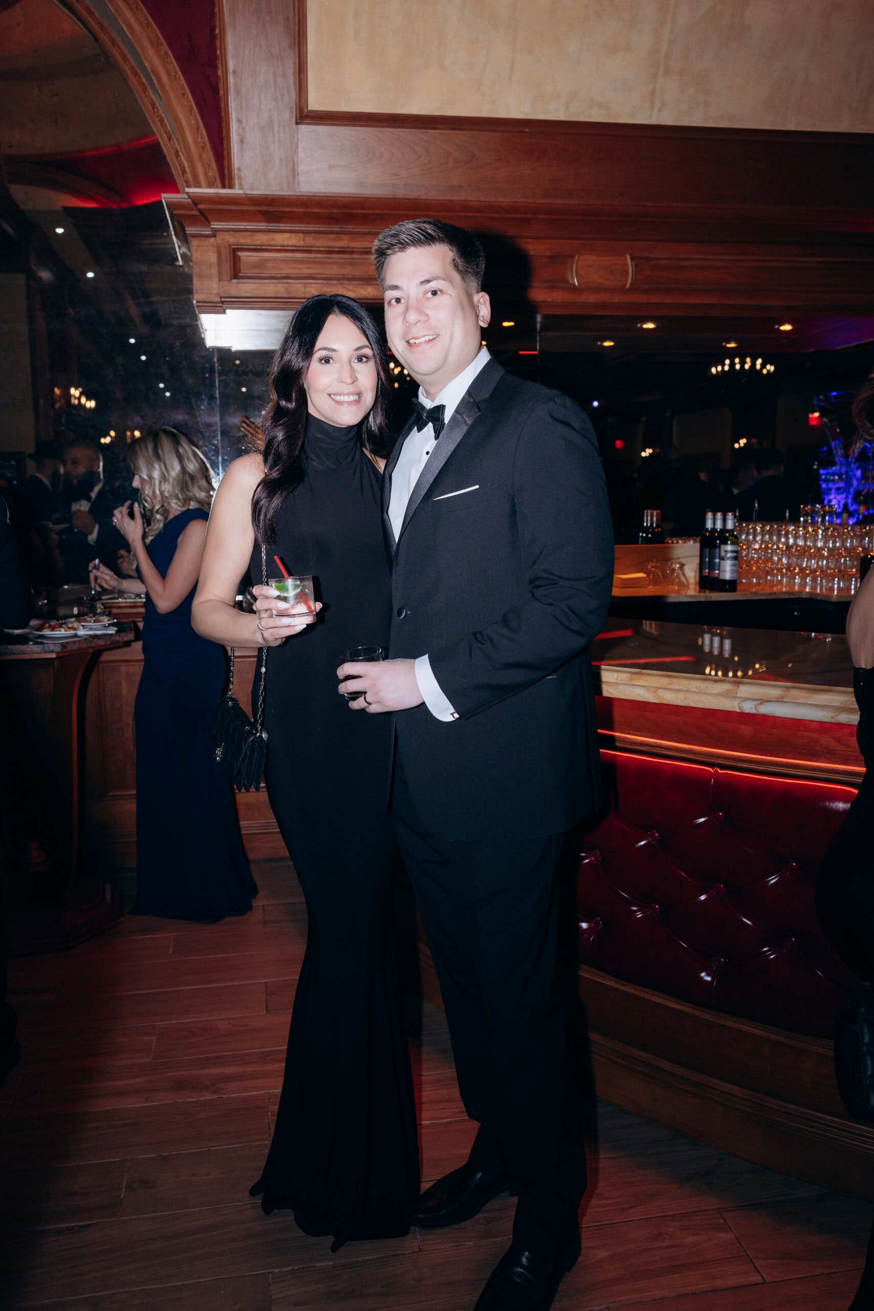 Man and woman dressed in black formal attire standing together in an elegant bar setting with wood paneling and a mirrored wall.