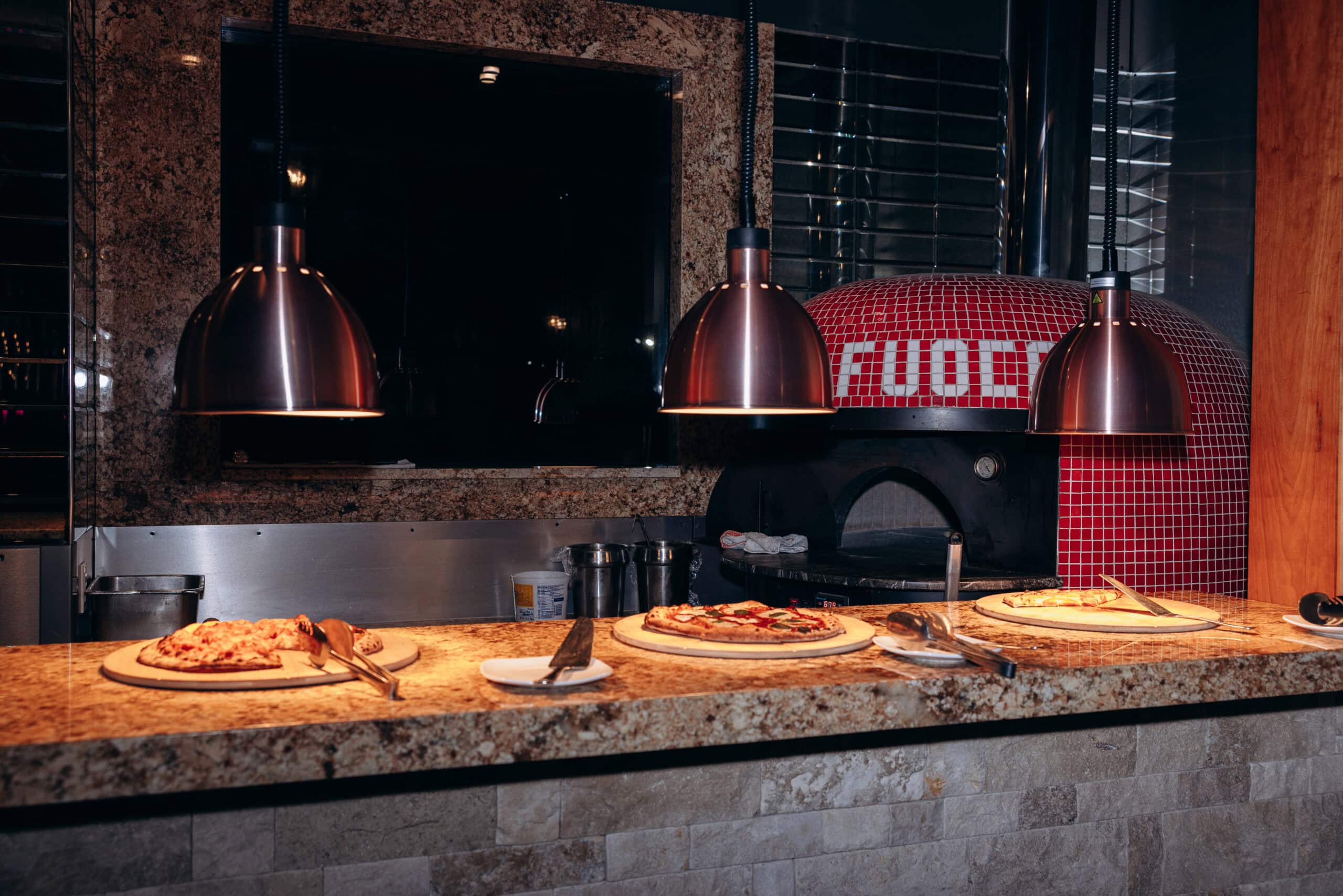 Three pizzas on wooden boards on a granite counter under copper pendant lights, with a red mosaic-tiled pizza oven in the background.