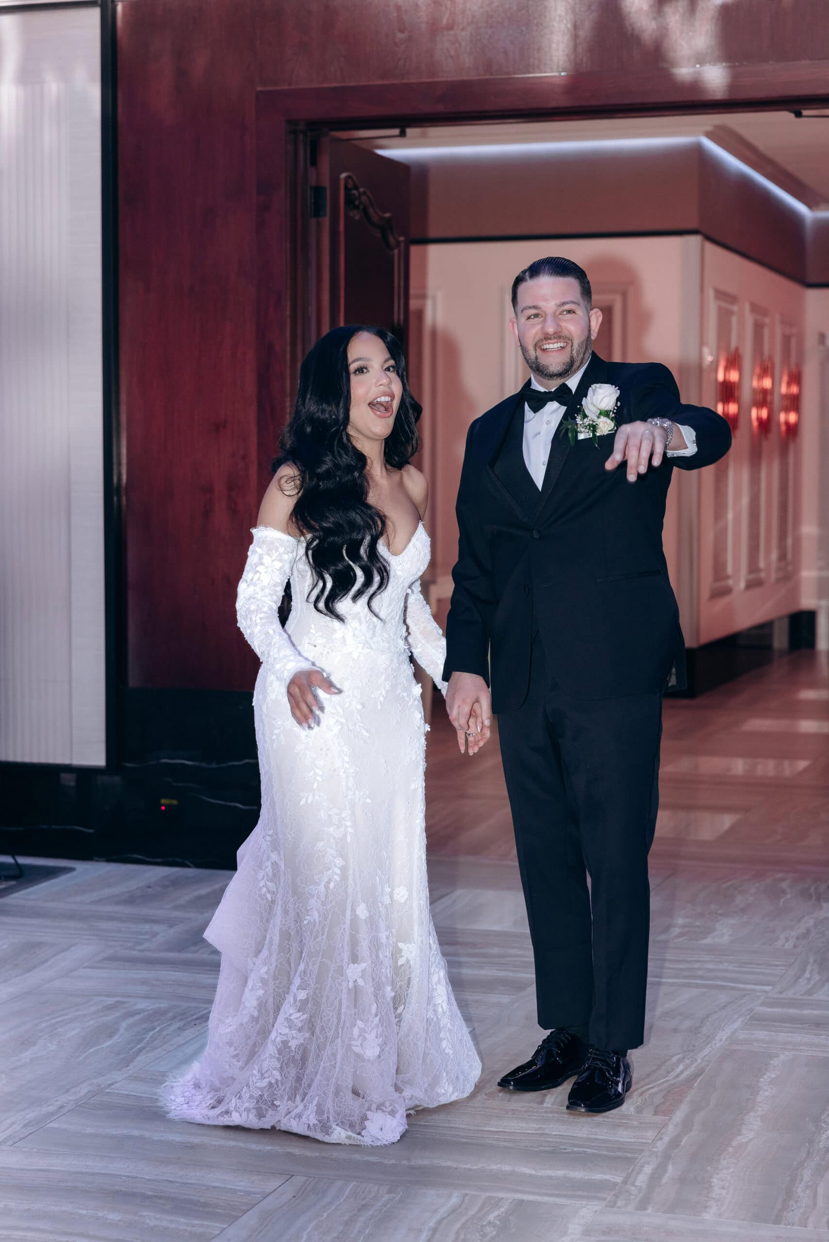 Bride in lace off-shoulder wedding dress and groom in black tuxedo holding hands, smiling and entering a venue with wood-paneled walls and ambient lighting.