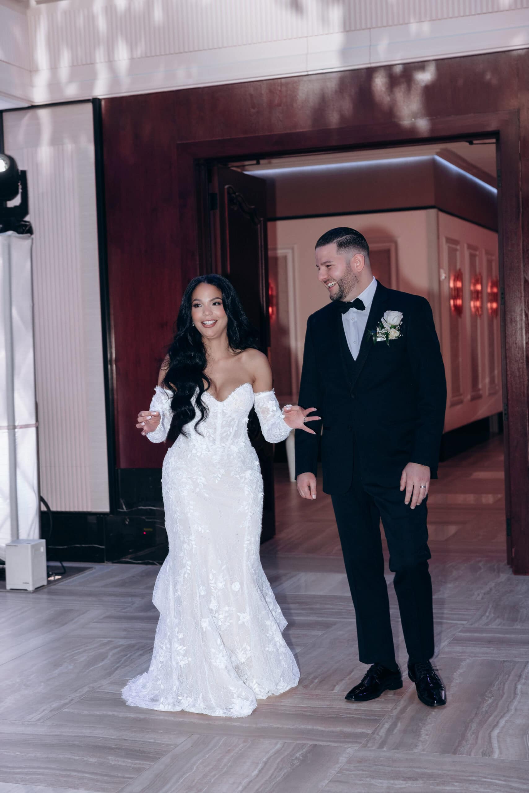 Bride in lace off-shoulder wedding dress and groom in black tuxedo walking into reception hall with wooden doors and illuminated exit sign above.