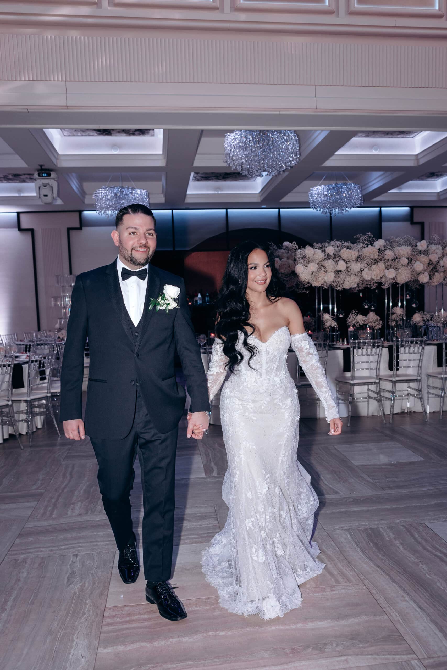 Bride and groom holding hands walking into a wedding reception with white floral centerpieces and purple ambient lighting at The Grand.