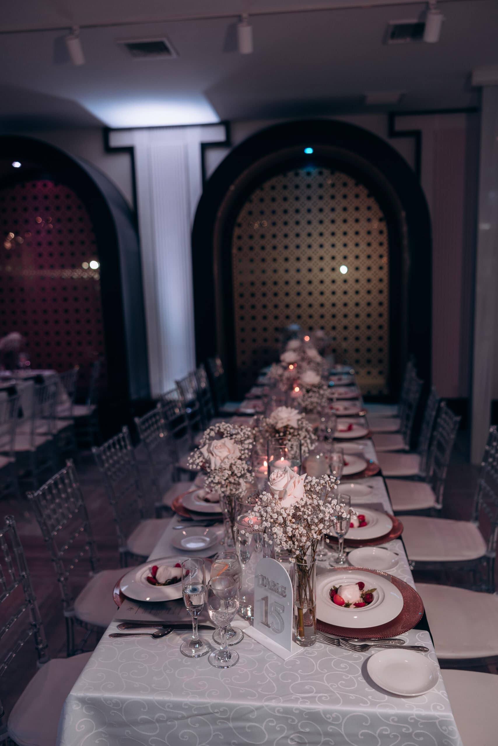 Long wedding reception table at The Grand with white roses and baby's breath floral arrangements, white patterned tablecloth, clear chairs, and soft purple ambient lighting.
