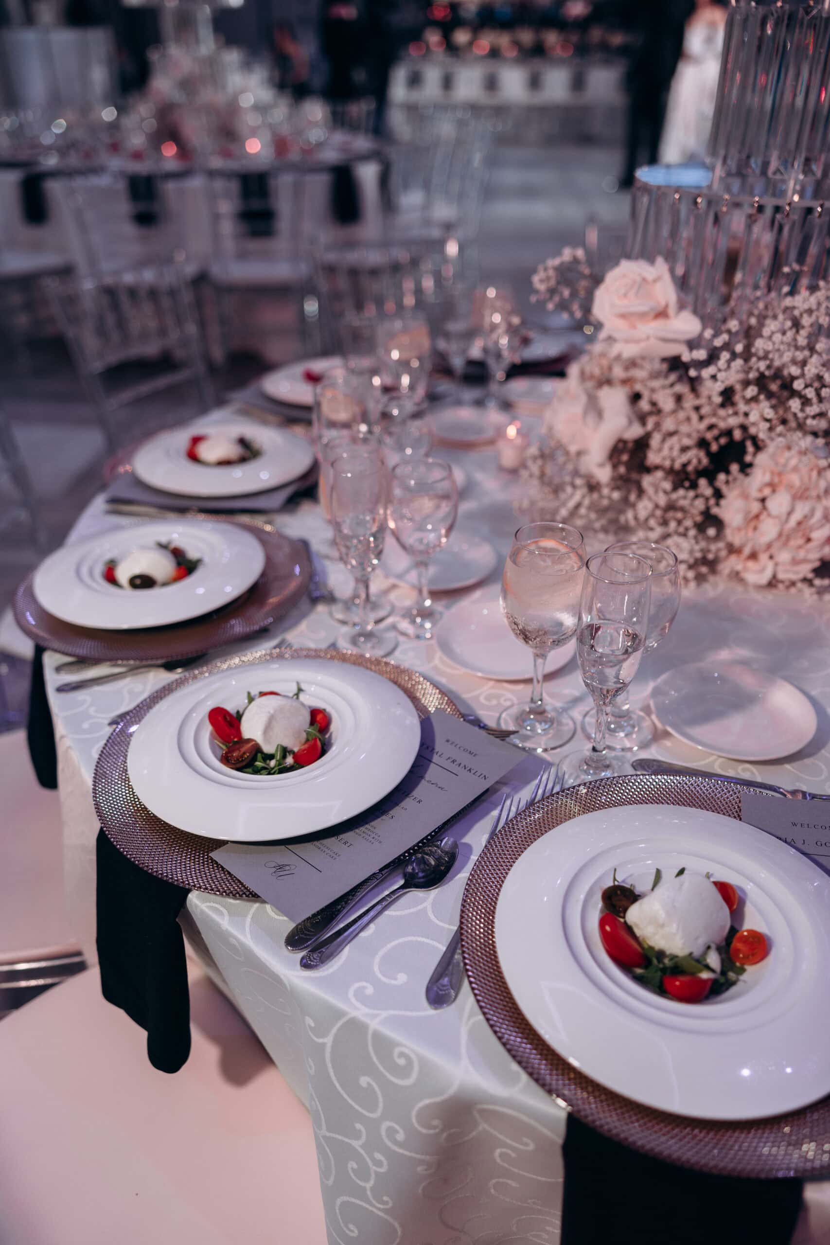 Close-up of a wedding table setting with white plates holding mozzarella and cherry tomato salads, crystal glasses, silver cutlery, and a white floral centerpiece with roses and baby's breath at The Grand.