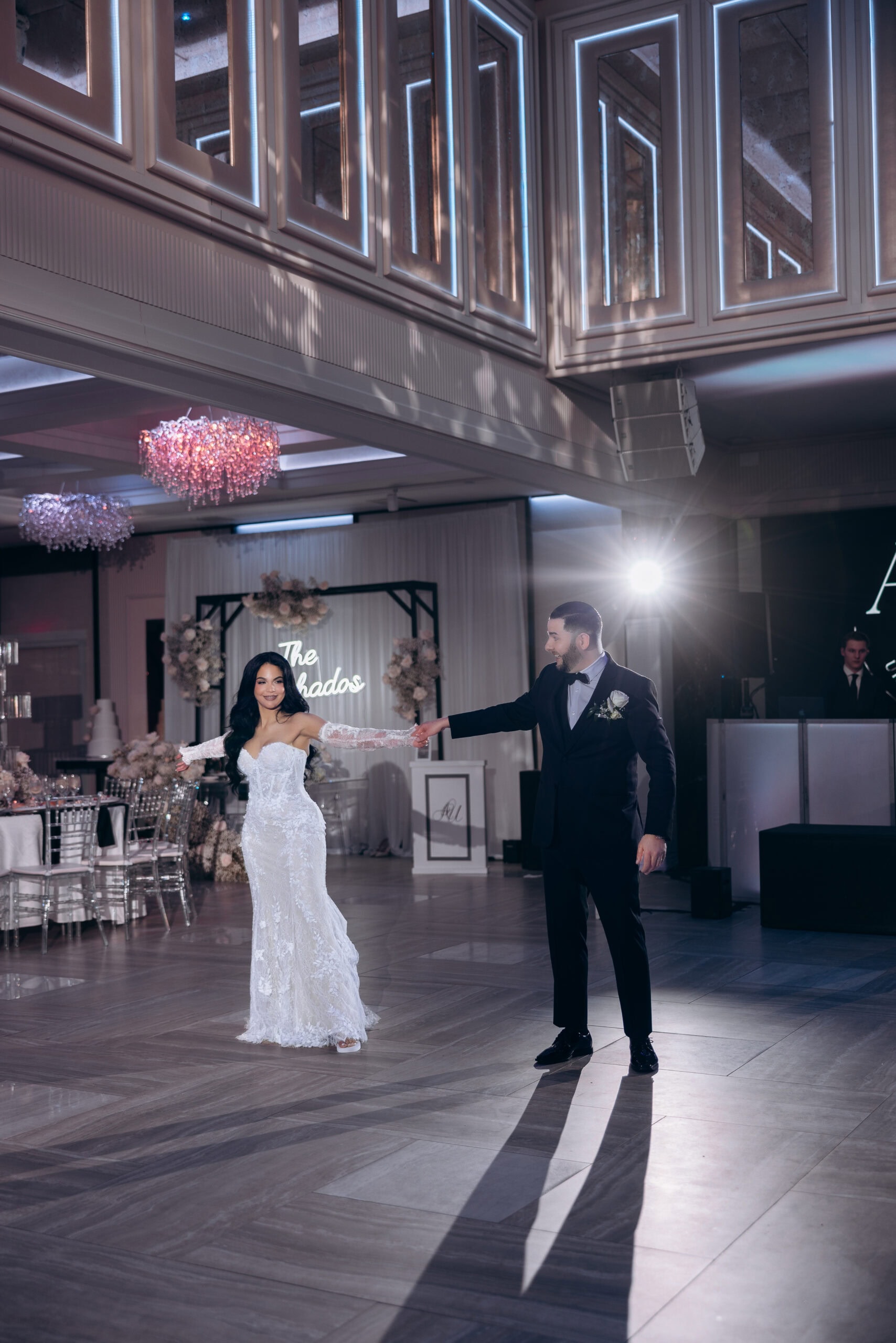 Bride and groom holding hands during their first dance in a softly lit ballroom with crystal chandeliers and floral arrangements at The Grand.