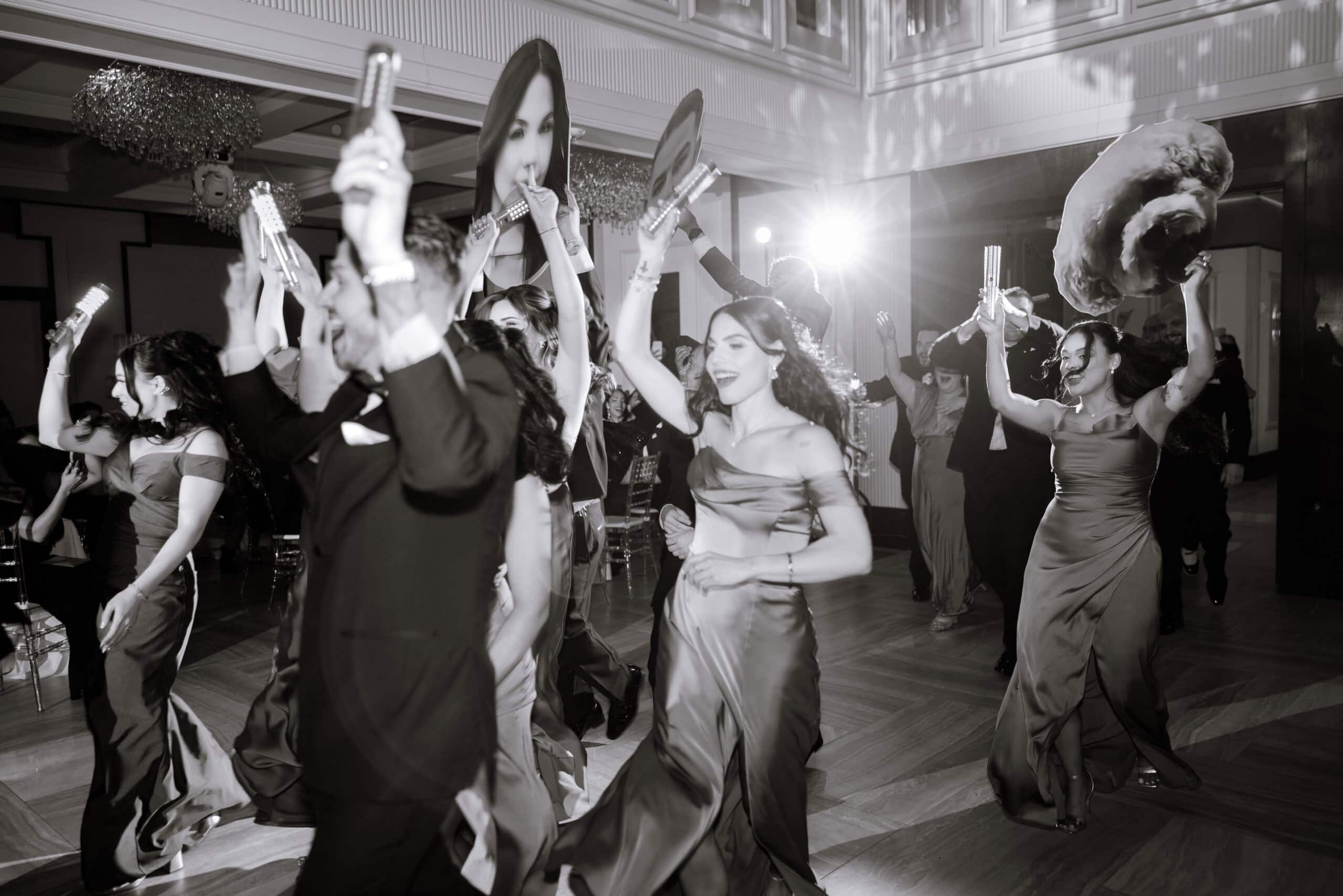 Wedding guests dancing energetically indoors, holding illuminated sticks and large playful face masks, dressed in formal taupe gowns and tuxedos under warm lighting.