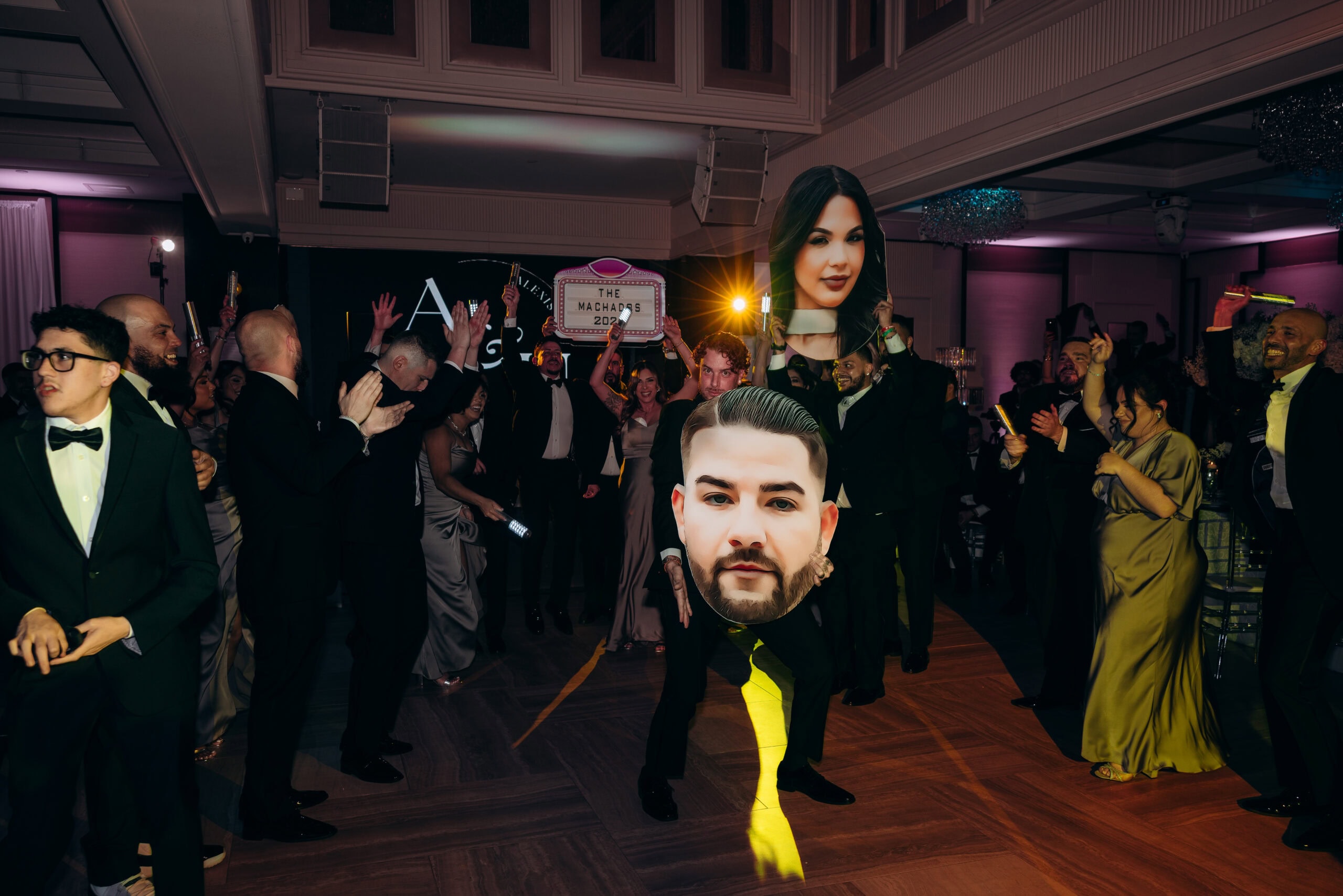 Wedding guests in formalwear at The Grand ballroom celebrating with illuminated sticks and oversized face cutouts, vibrant lighting and joyful atmosphere.
