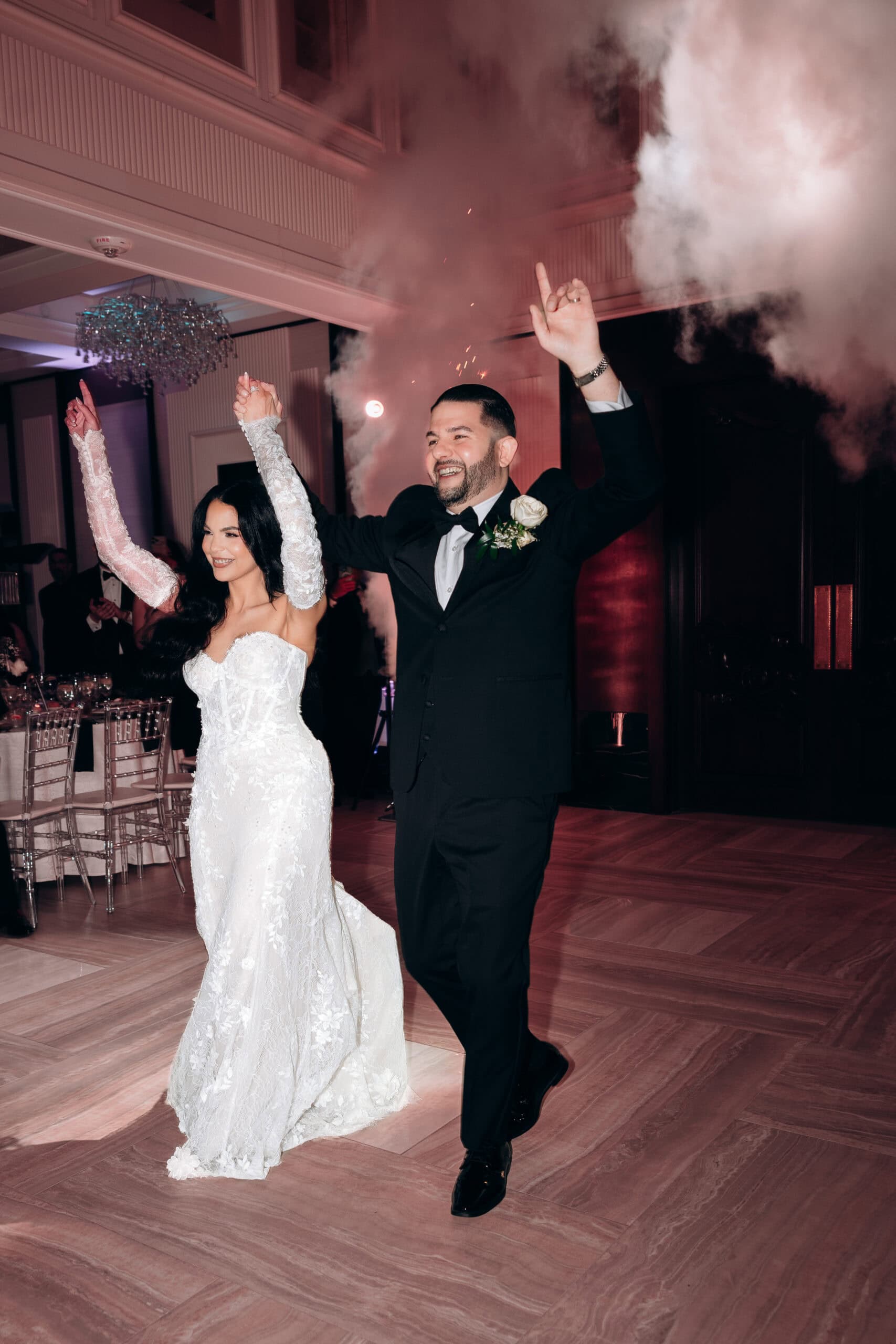 Bride in lace wedding dress and groom in black tuxedo entering reception with raised hands, surrounded by smoke and sparks, at a luxury venue.