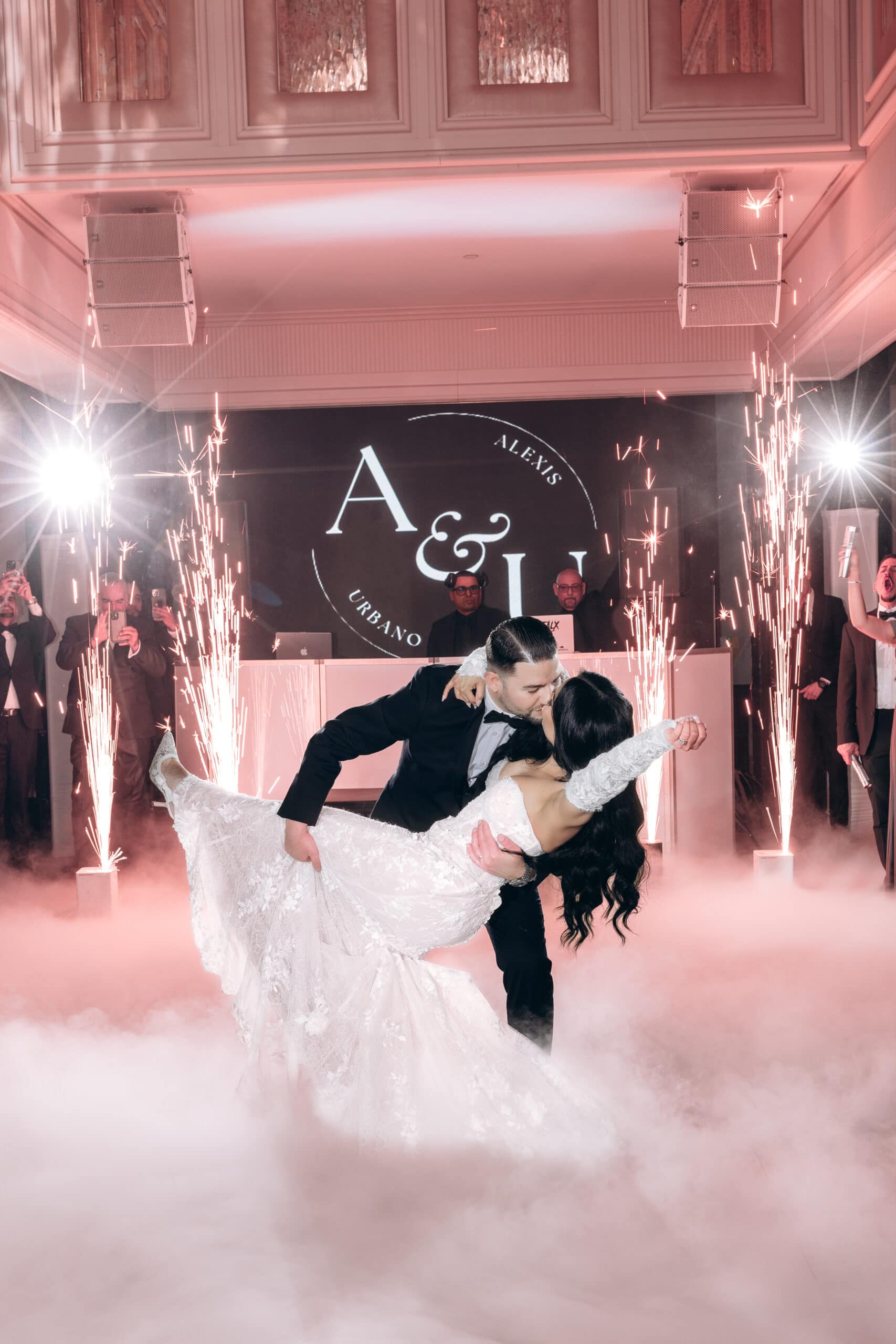Groom dipping bride in lace gown on foggy dance floor with sparklers and illuminated monogram backdrop at The Grand wedding venue.