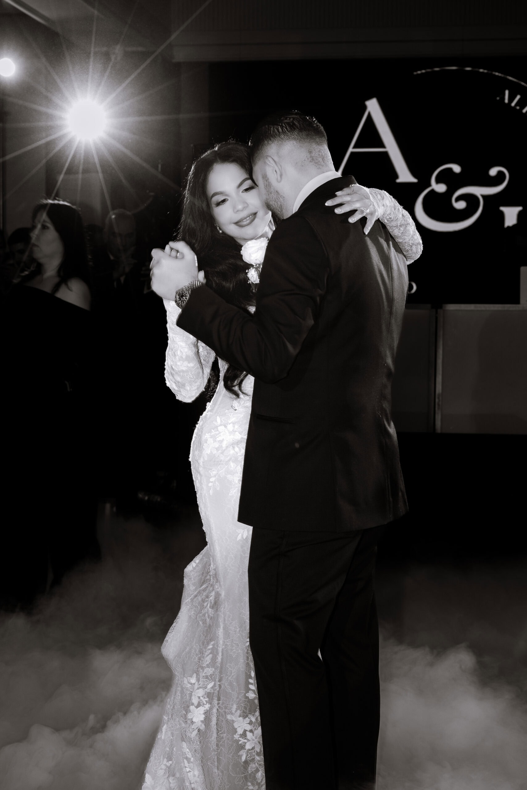 Bride and groom dancing closely during their wedding first dance, surrounded by low fog and illuminated by a bright backlight, with guests in the background.