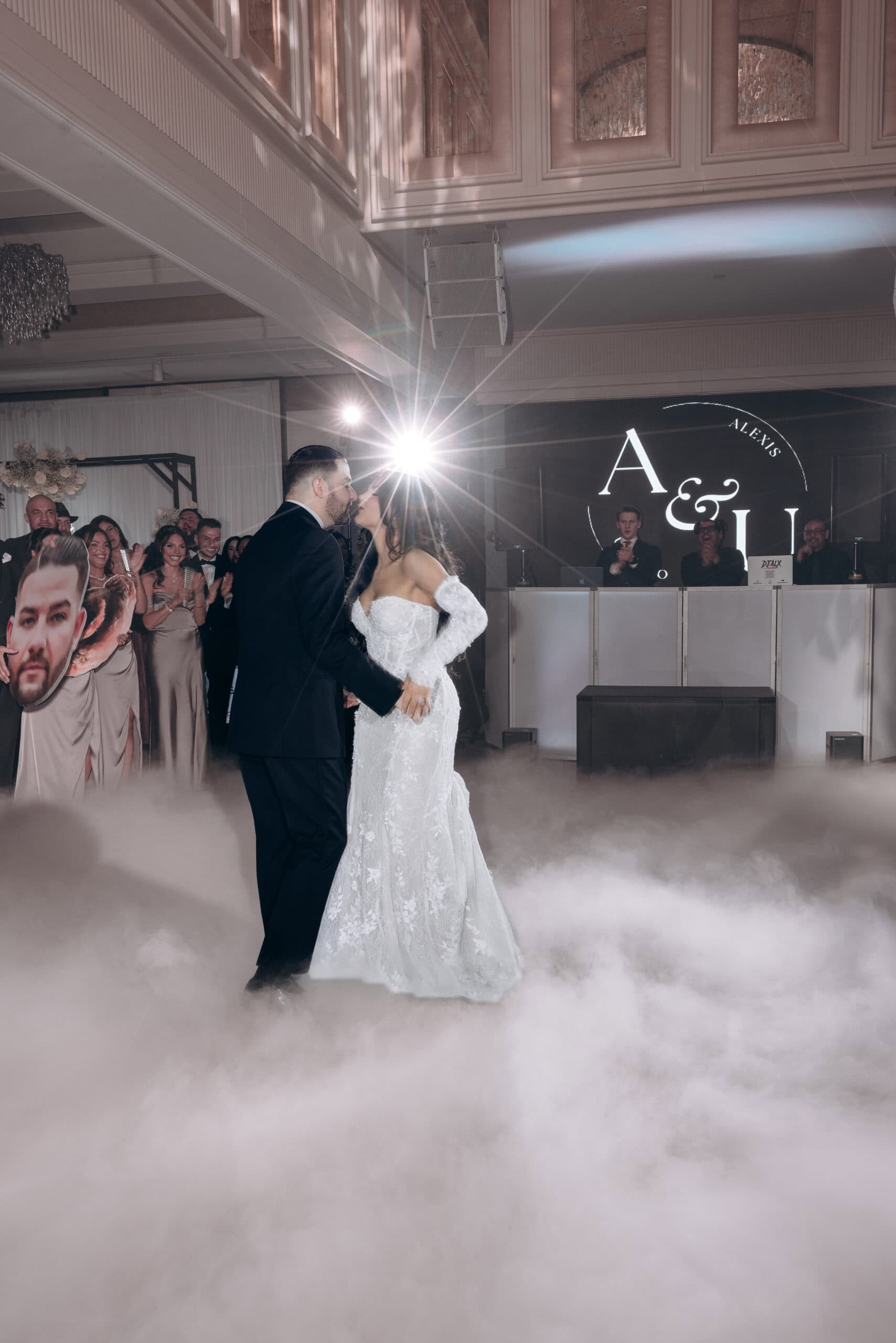 Bride and groom dancing in a fog-filled ballroom under a bright spotlight, guests applauding nearby, with a DJ booth and monogrammed backdrop at The Grand.