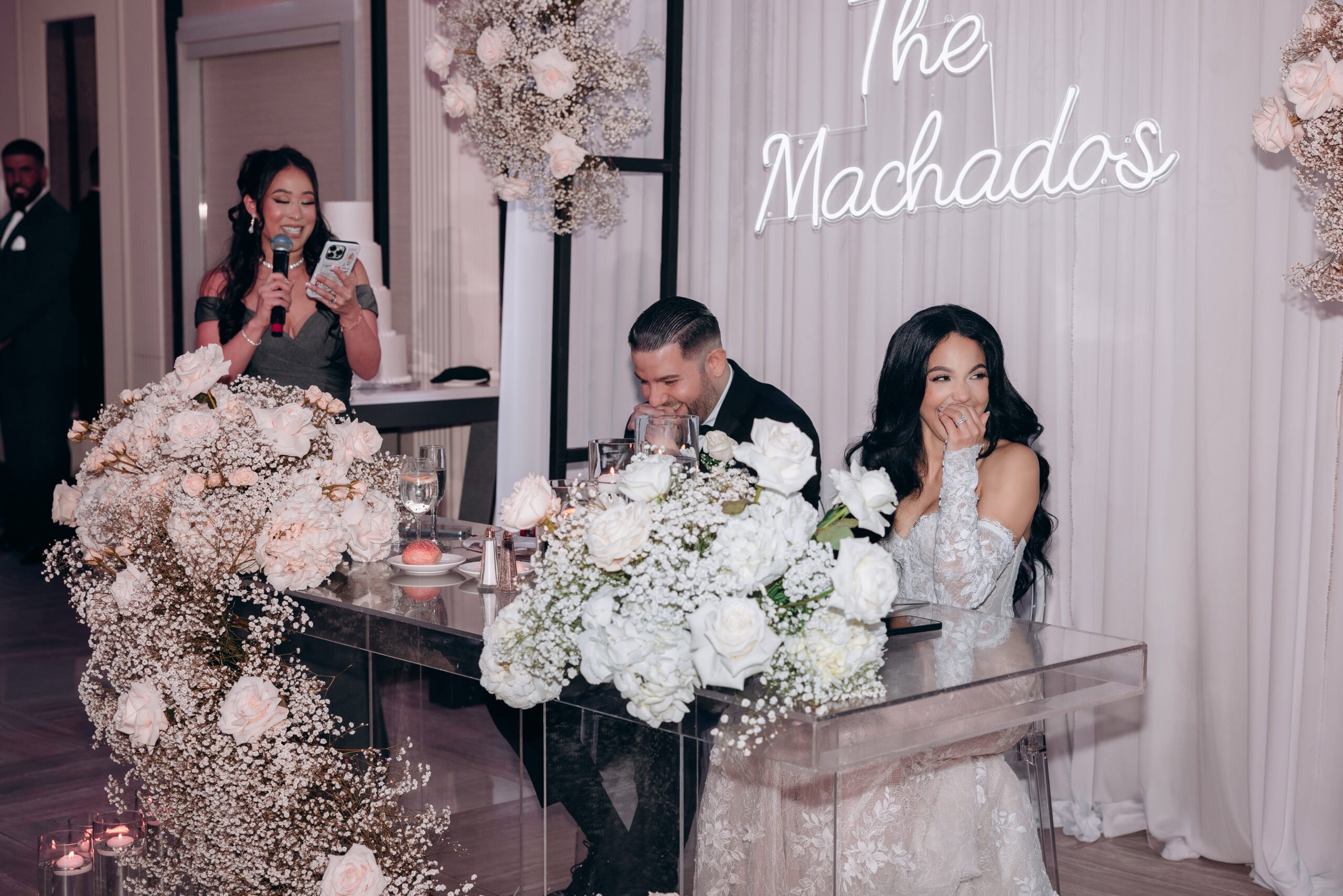 Bride and groom seated at a glass table adorned with white roses and baby's breath, listening to a woman delivering a speech with a microphone and phone, illuminated neon sign reading 'The Machados' in the background.