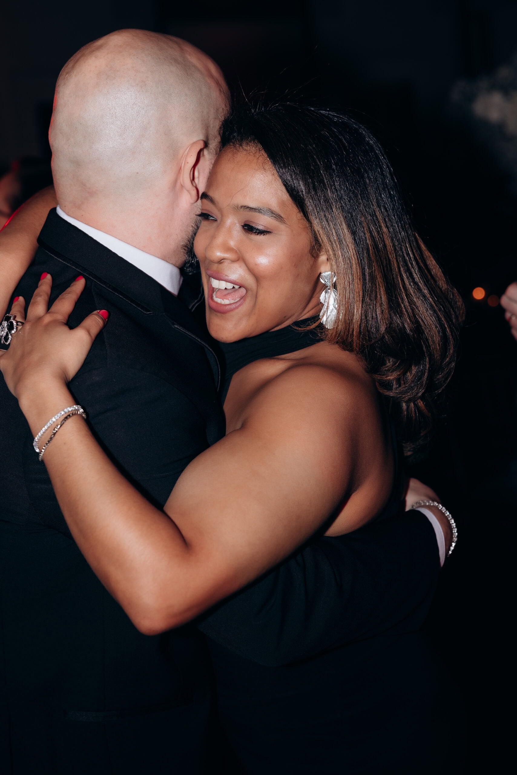 Woman with silver statement earrings and manicured nails embraces a bald man in a black suit during a dance at an evening event.