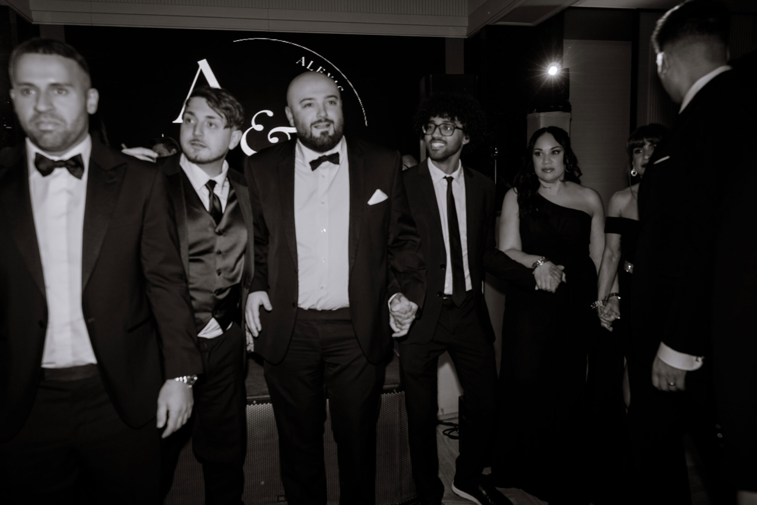 Group of elegantly dressed individuals in black-tie attire holding hands in a dimly lit indoor setting with a circular illuminated sign in the background.