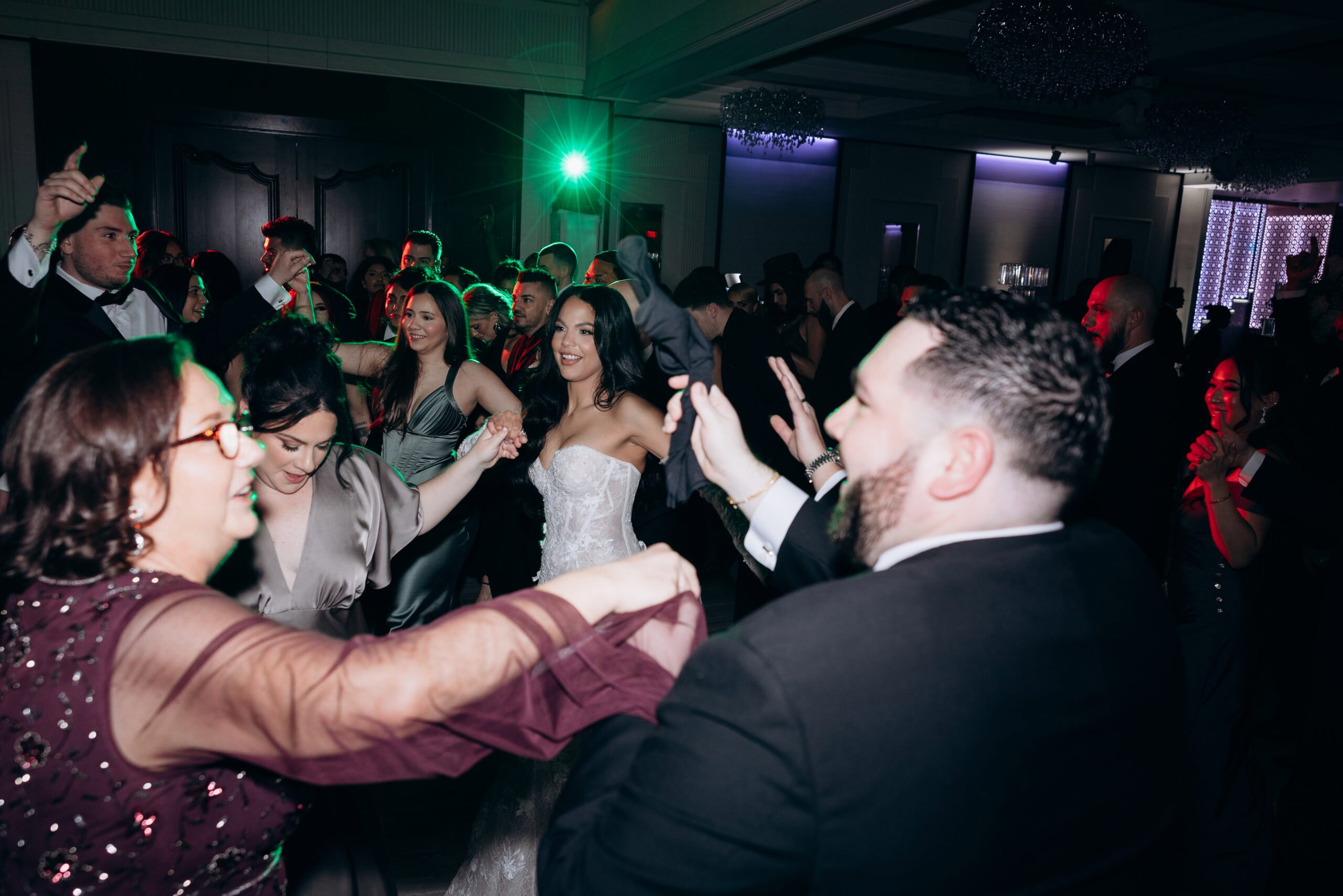 Wedding guests dancing in a dimly lit room with green and red accent lighting, featuring a bride in a lace gown and guests in formalwear.